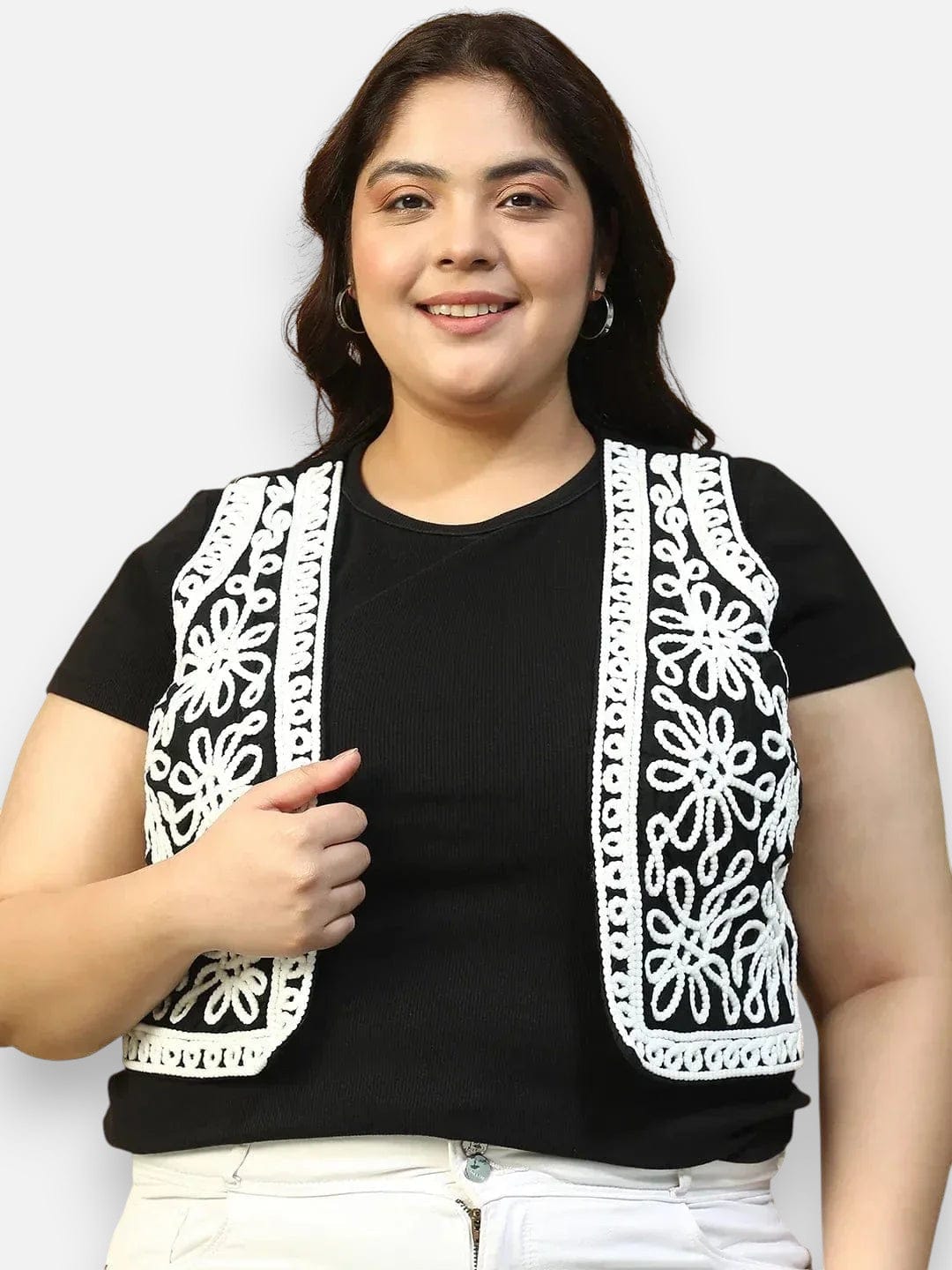 A woman with dark hair is smiling and wearing a black top with a white floral pattern on the vest.