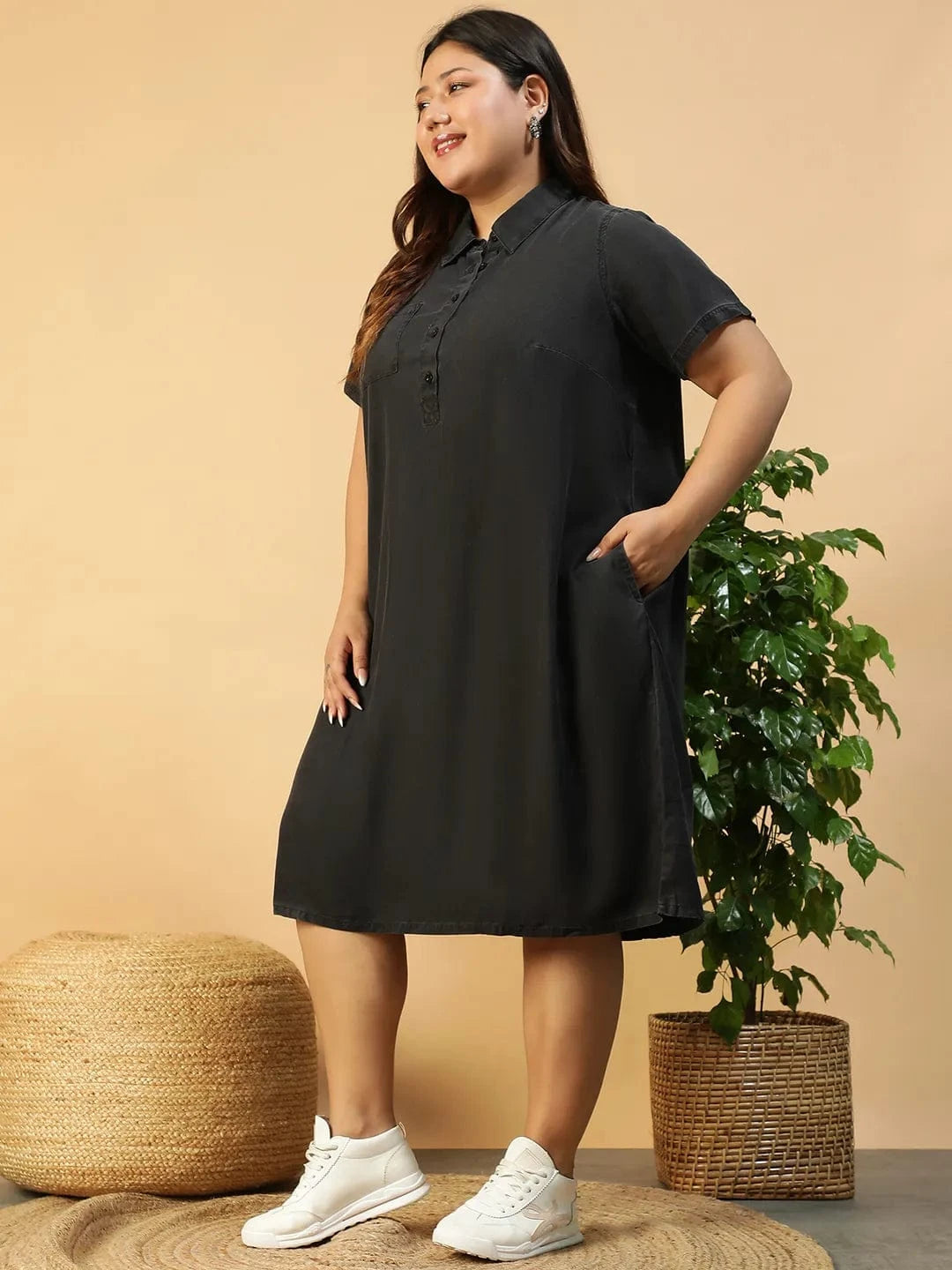 A woman wearing a black dress with short sleeves and a collar, posing with her hand on her hip and smiling.