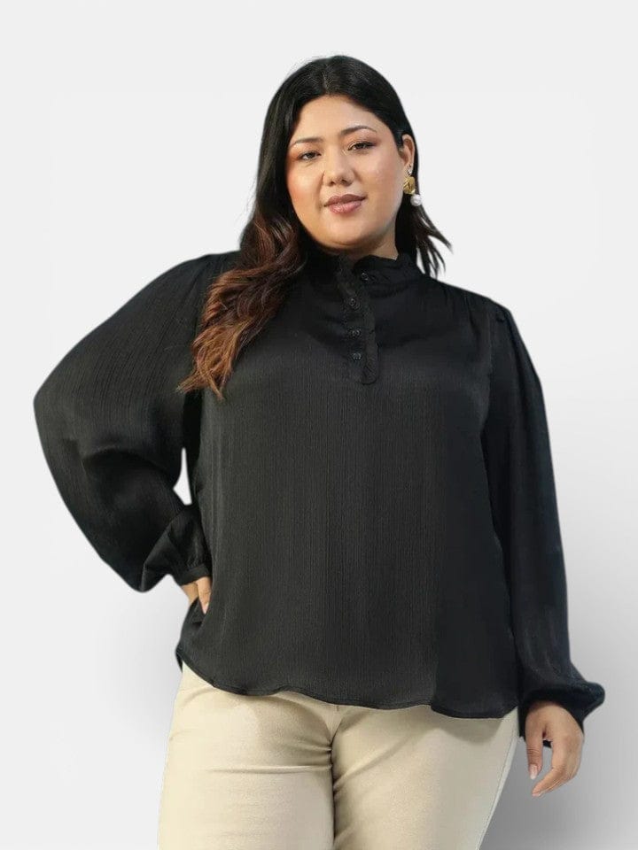 A woman with long dark hair wearing a black blouse and beige pants stands against a white background, smiling at the camera.