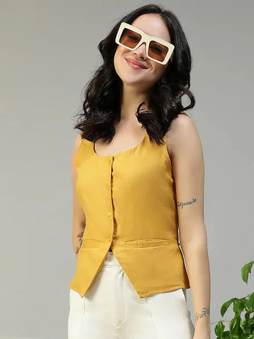 Woman in mustard top and white pants standing in a room with a plant and chair.
