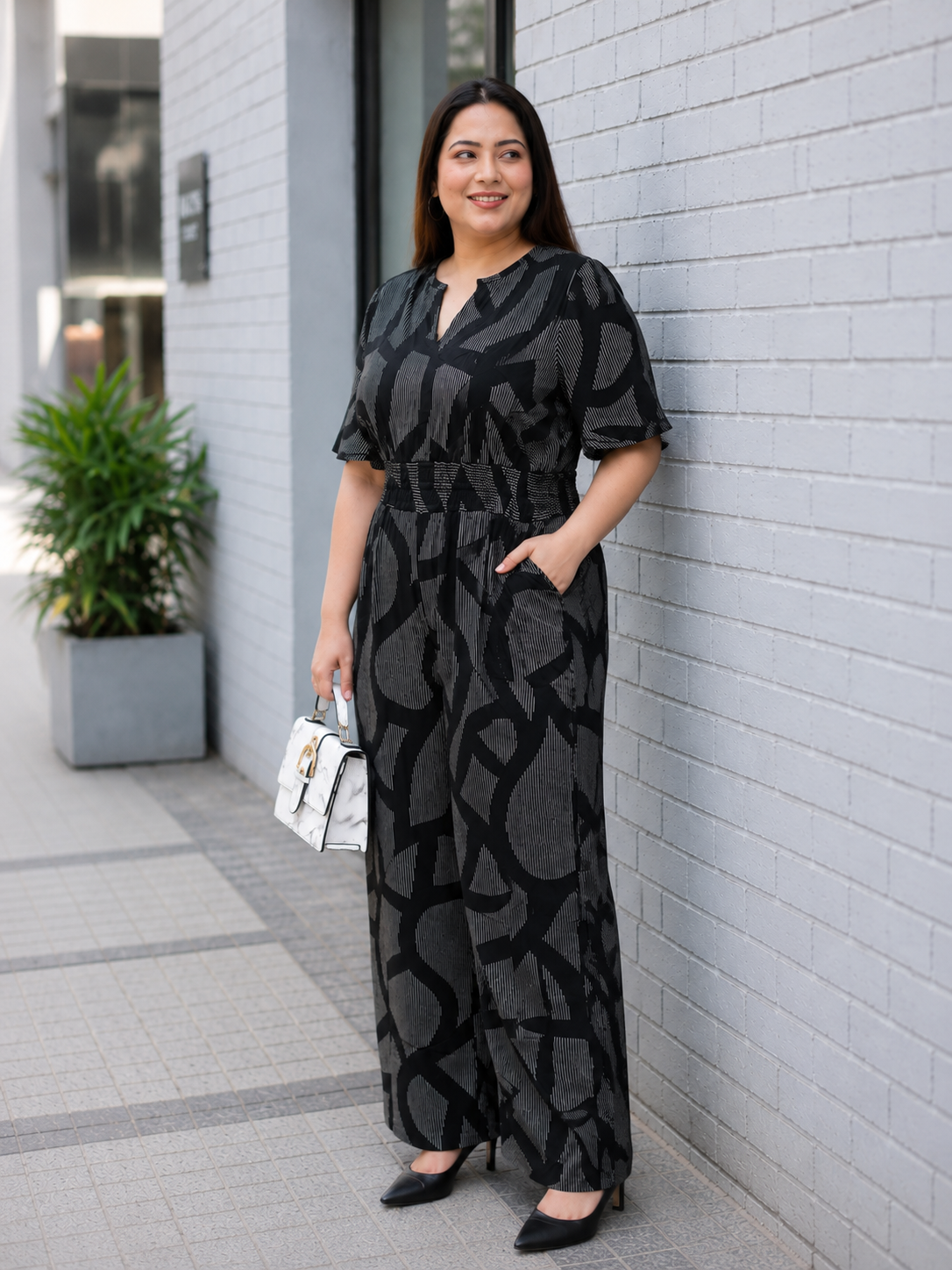 A woman wearing a black jumpsuit with a gray pattern stands in front of a white brick wall, smiling and holding a white handbag.