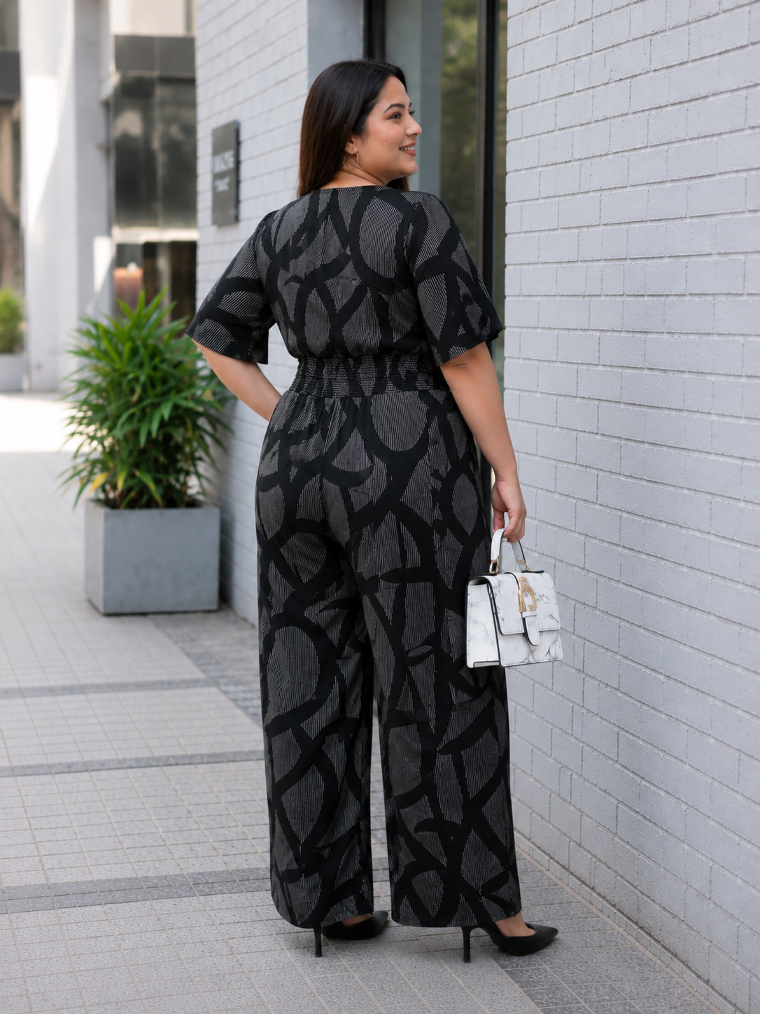 A woman wearing a black jumpsuit with a black and gray pattern stands in front of a white brick wall, smiling and holding a white handbag.
