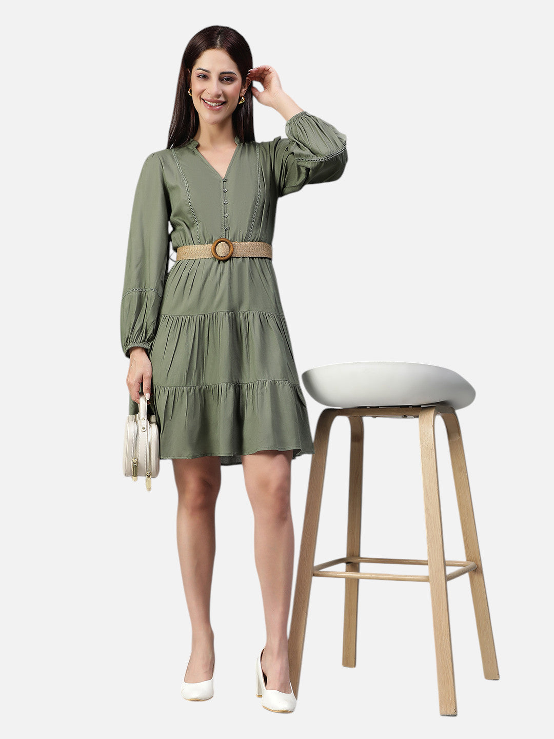 Woman in olive green belted dress with long sleeves, holding a handbag, standing by a stool