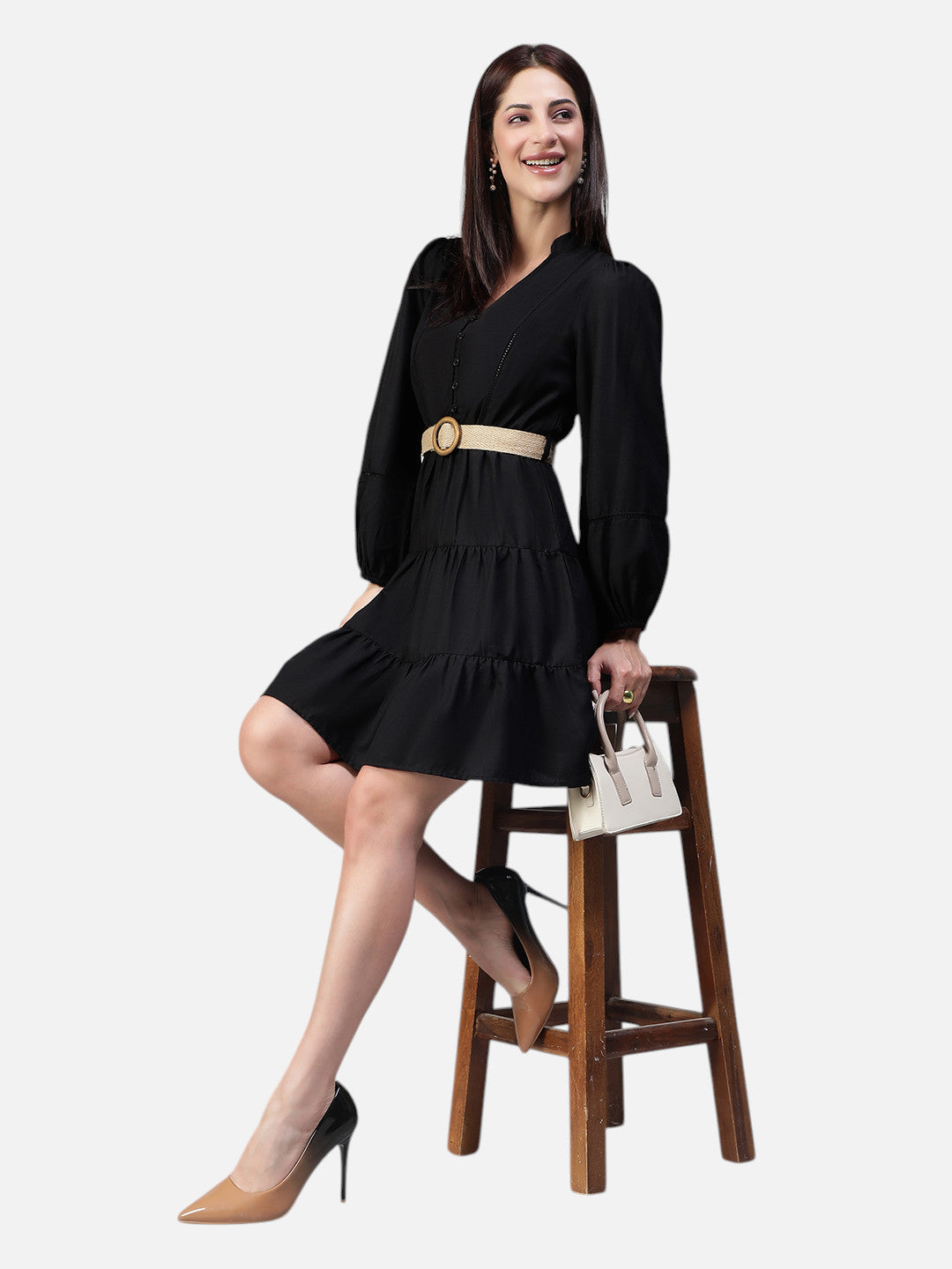 Woman in black long sleeve belted mini dress, sitting on a stool with beige handbag
