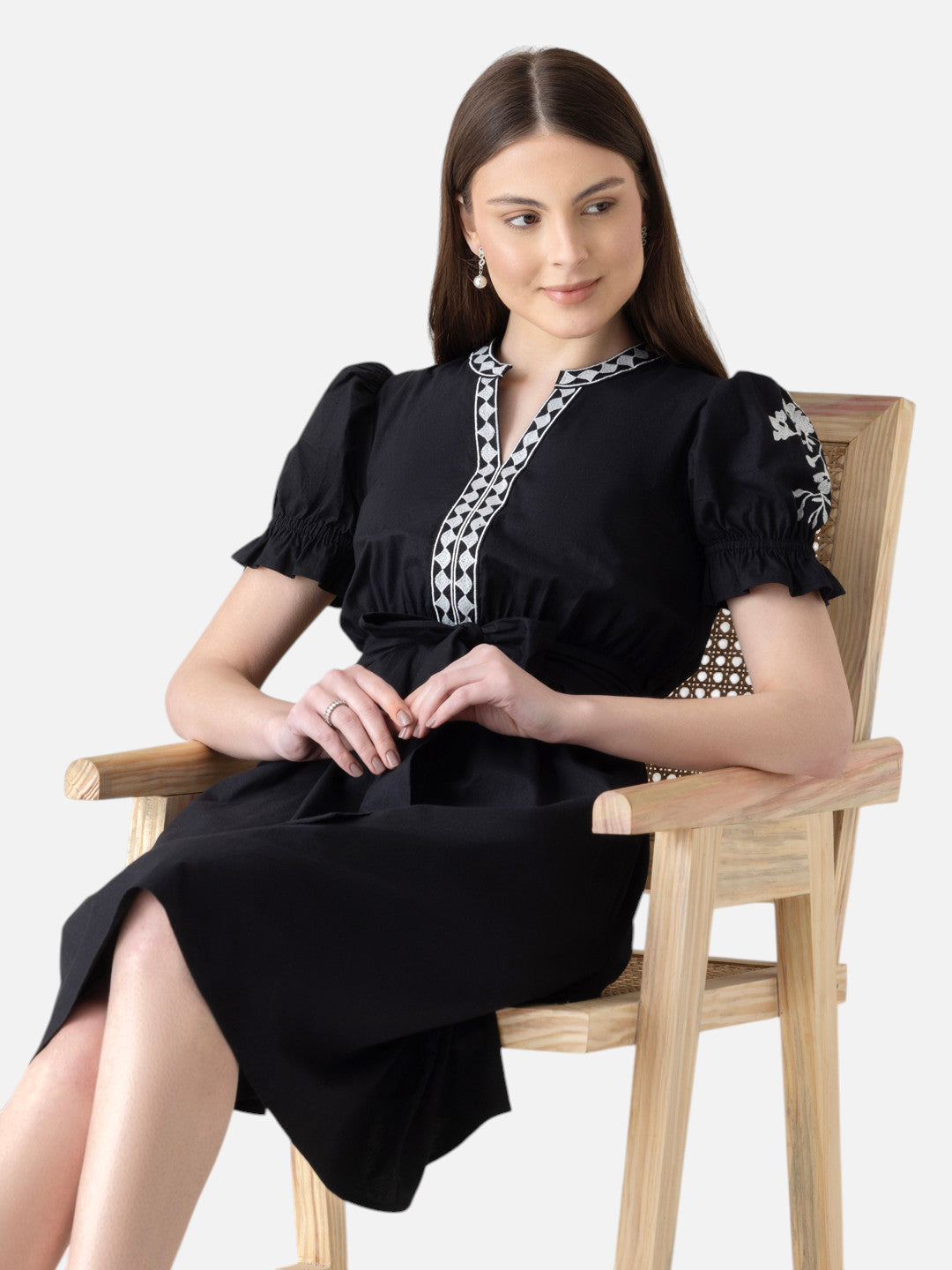 Woman in black knee-length dress with white embroidery sitting on wooden chair