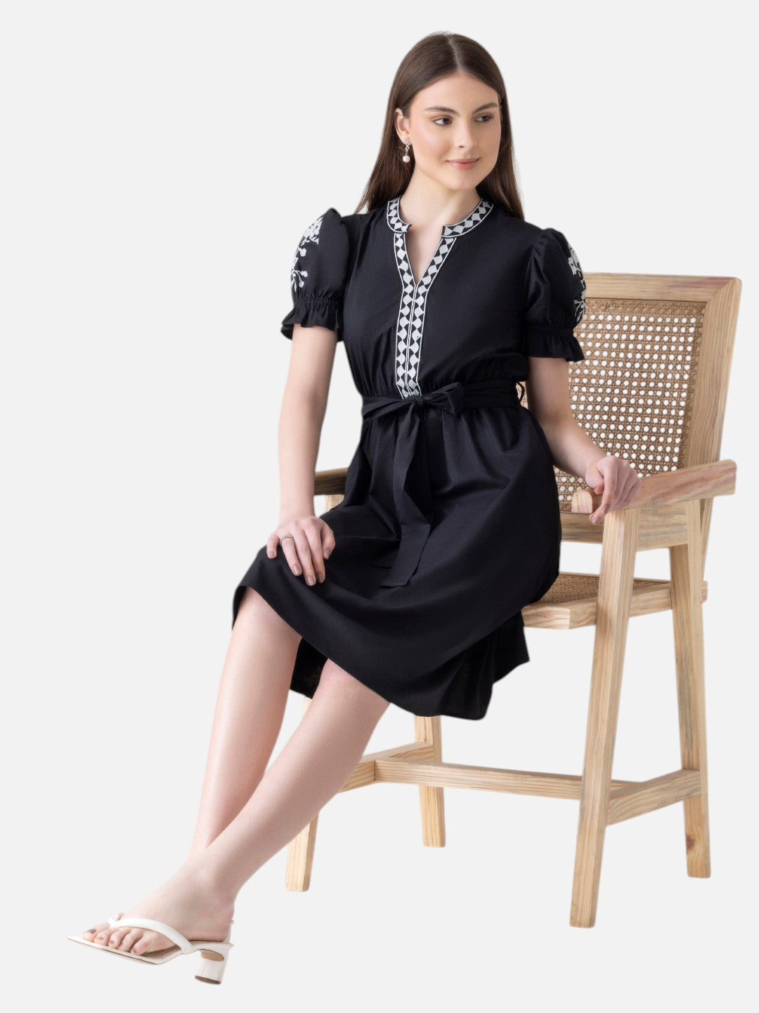 Woman sitting on chair wearing black knee-length dress with white embroidery and short puff sleeves