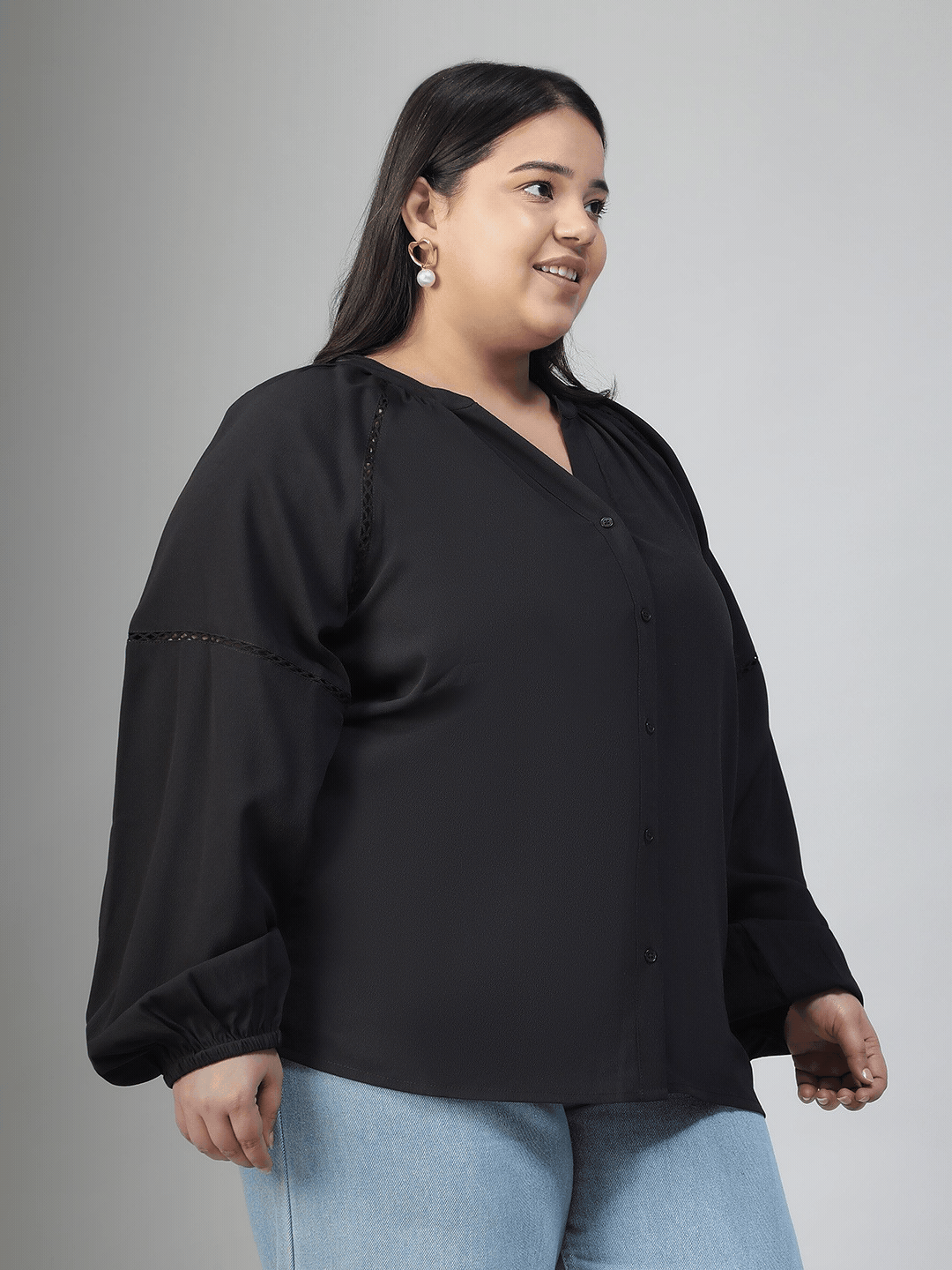 A woman with long dark hair is smiling and posing for the camera. She is wearing a black blouse with long sleeves and blue jeans.