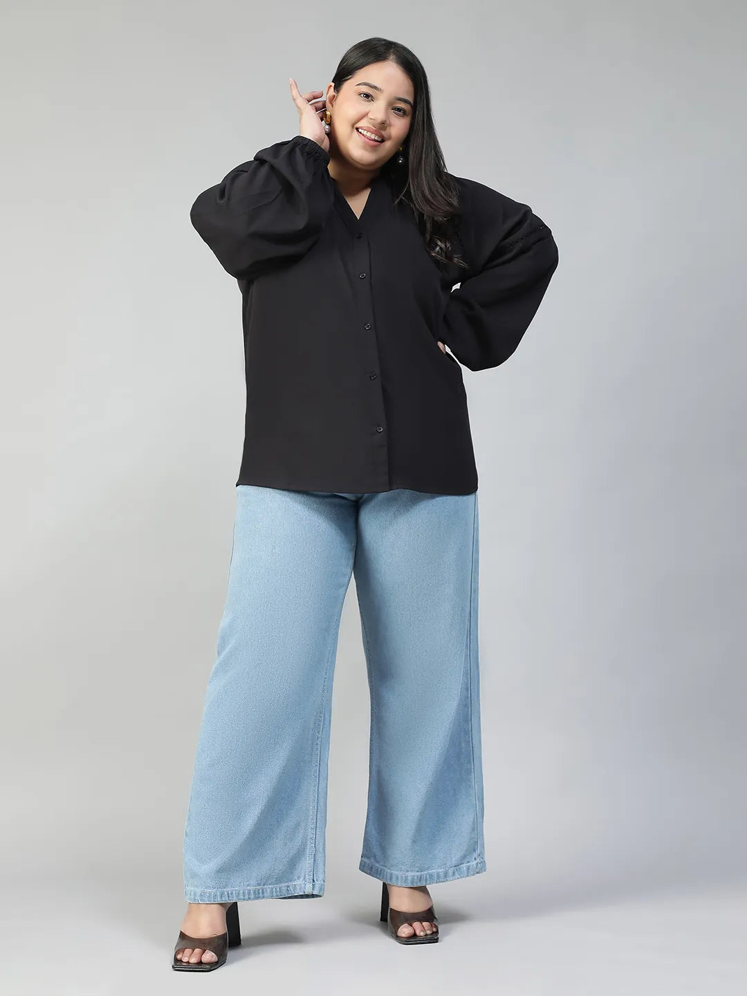 A woman with long dark hair is smiling and posing for the camera. She is wearing a black blouse with long sleeves and blue jeans.