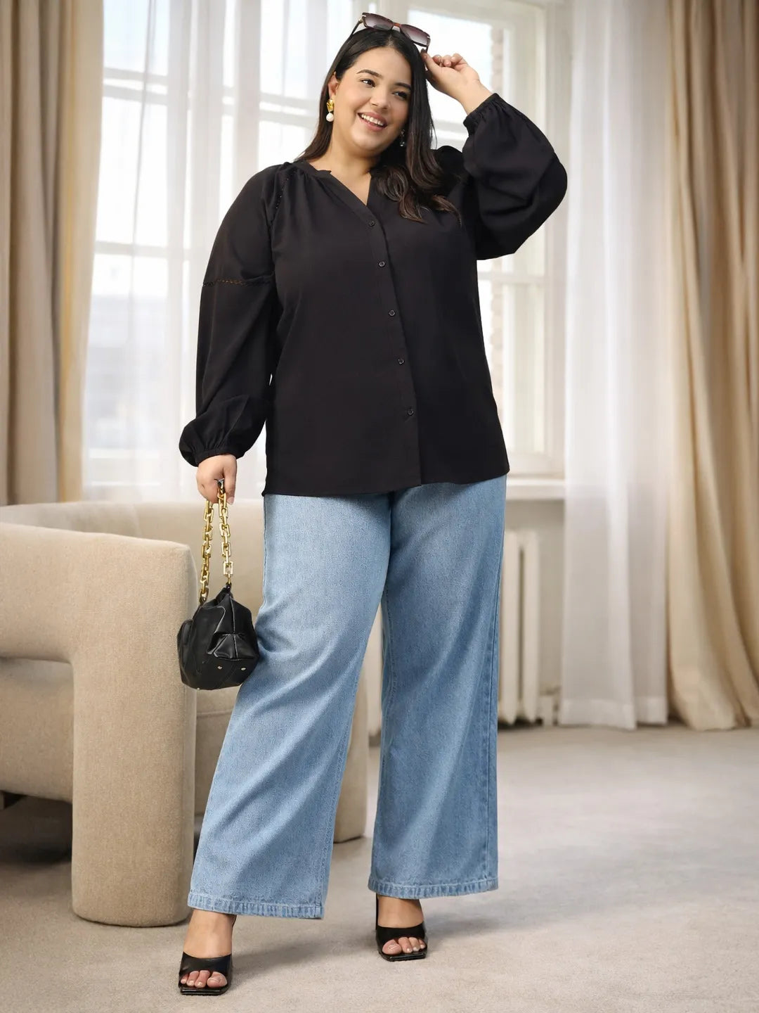 A woman with long dark hair is smiling and posing for the camera. She is wearing a black blouse with long sleeves and blue jeans.