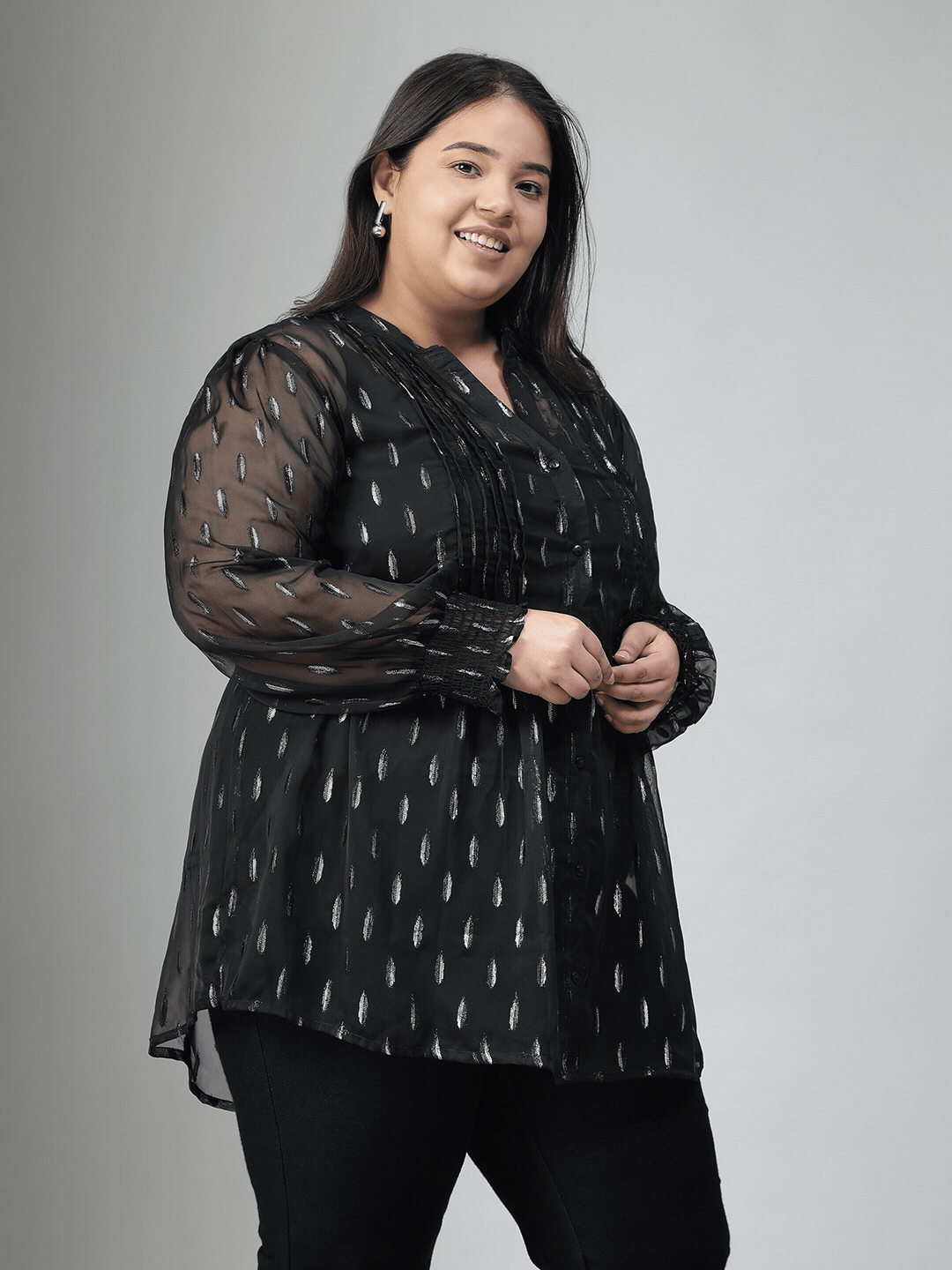 A woman with long dark hair is smiling and posing for the camera. She is wearing a black blouse with silver dots and has her hand on her hip.