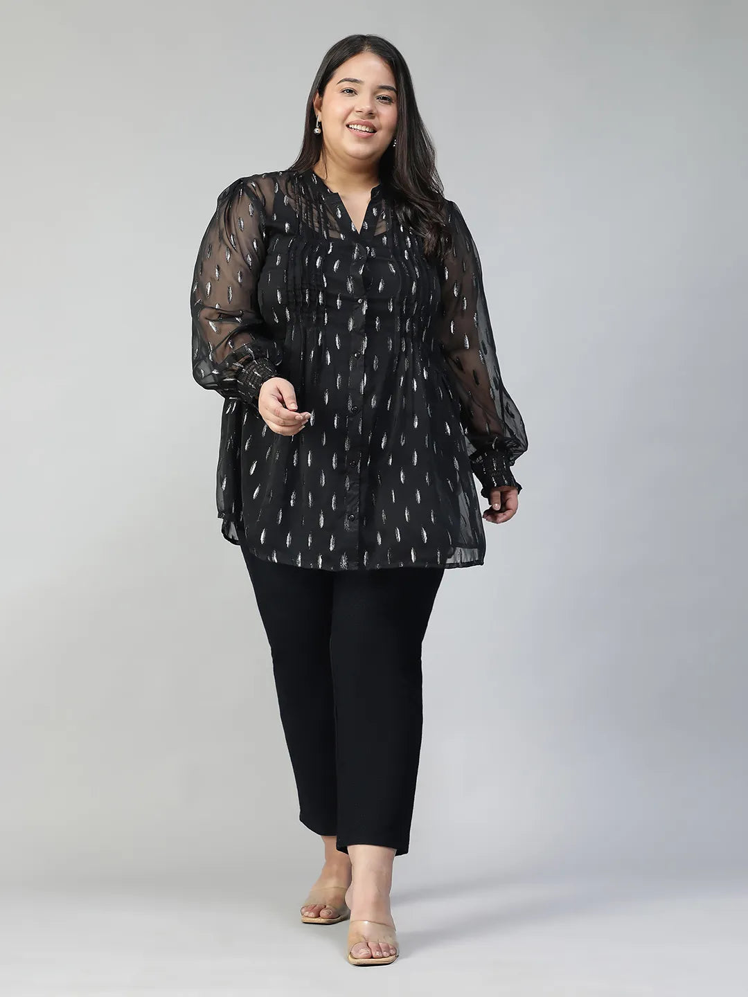 A woman with long dark hair is smiling and posing for the camera. She is wearing a black blouse with silver dots and has her hand on her hip.
