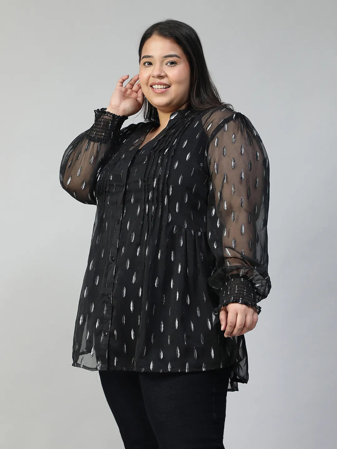 A woman with long dark hair is smiling and posing for the camera. She is wearing a black blouse with silver dots and has her hand on her hip.