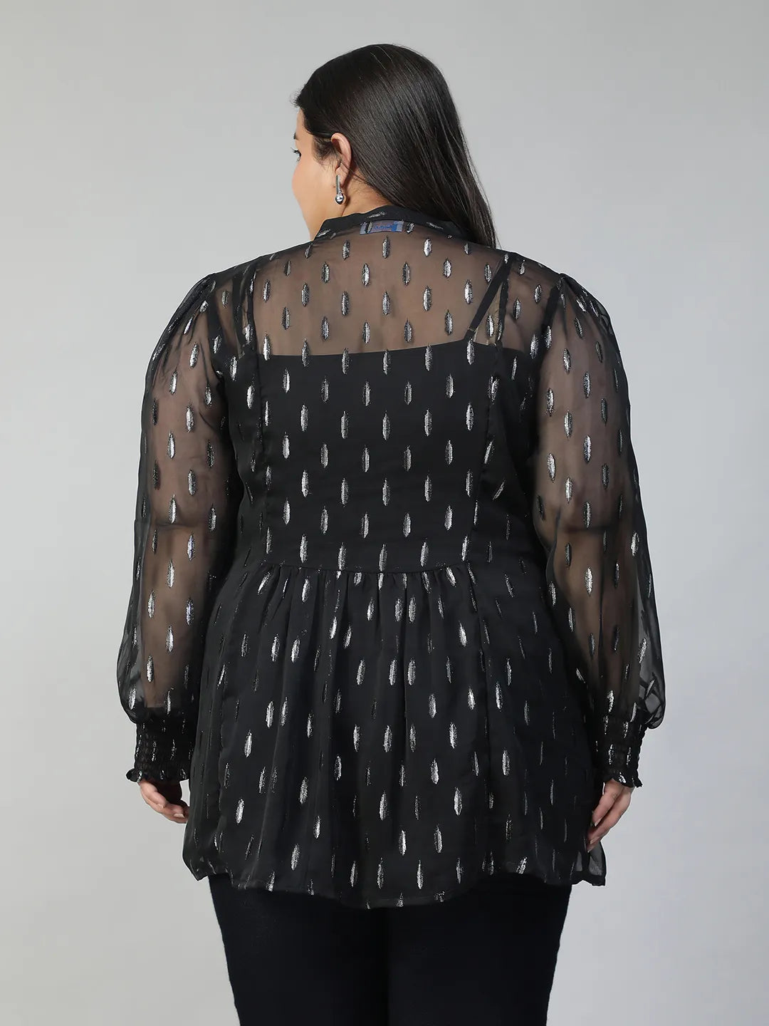 A woman with long dark hair is smiling and posing for the camera. She is wearing a black blouse with silver dots and has her hand on her hip.