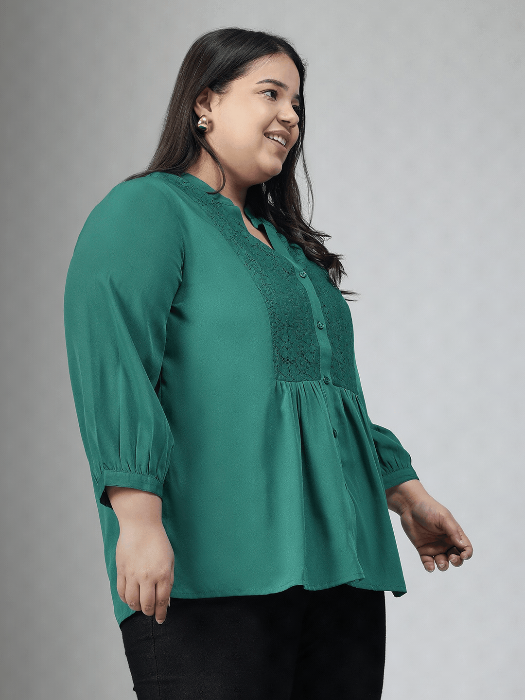 A smiling woman wearing a green blouse with lace detailing and puffed sleeves, posing confidently with her hand on her hip.