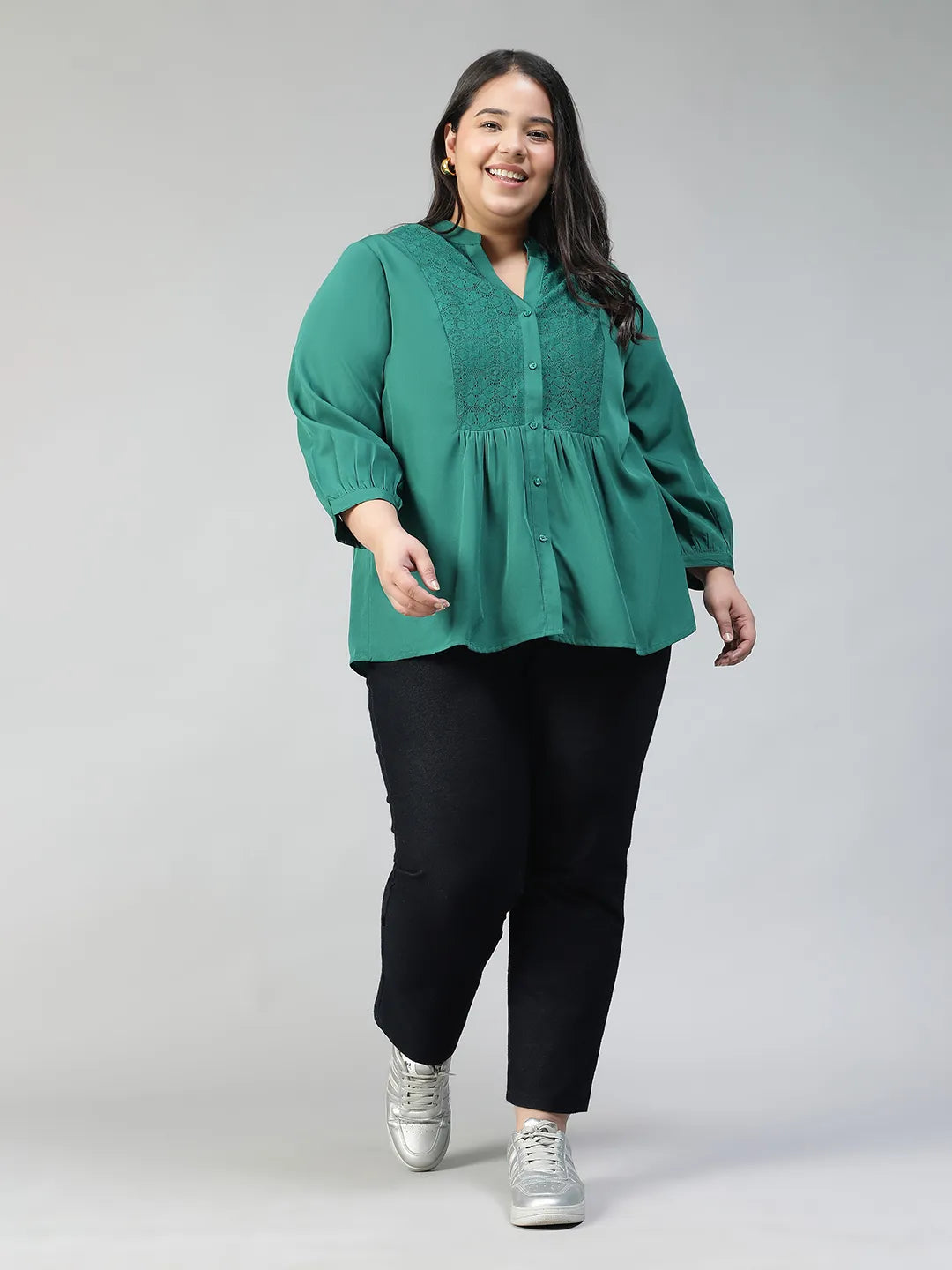 A smiling woman wearing a green blouse with lace detailing and puffed sleeves, posing confidently with her hand on her hip.