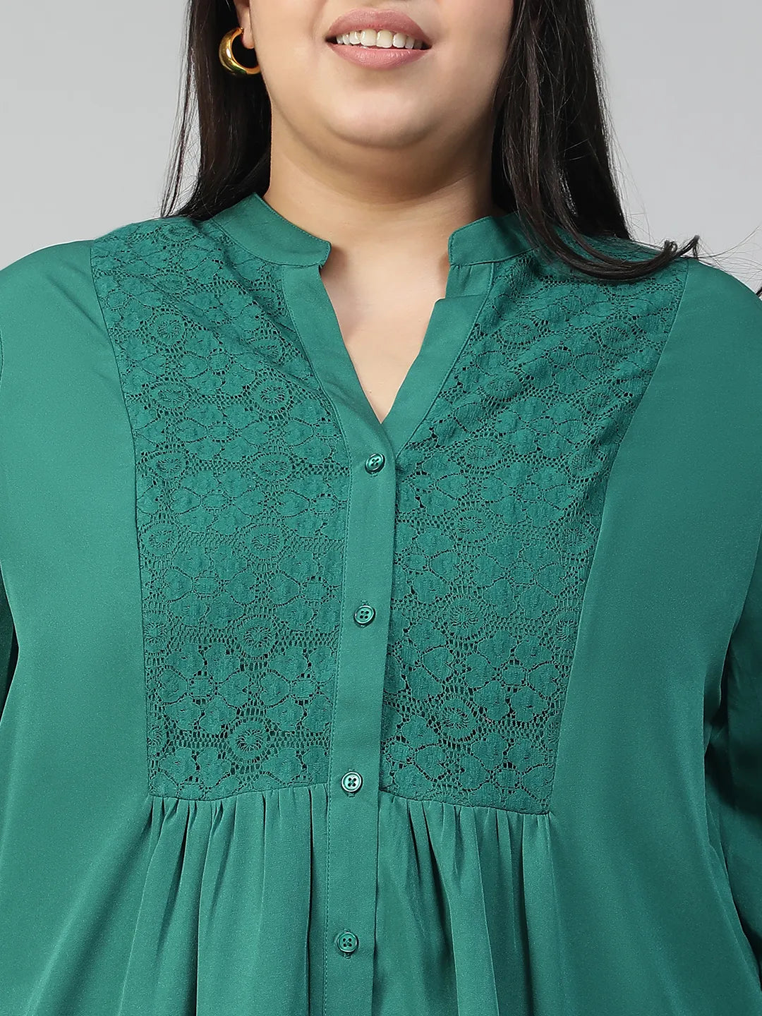 A smiling woman wearing a green blouse with lace detailing and puffed sleeves, posing confidently with her hand on her hip.
