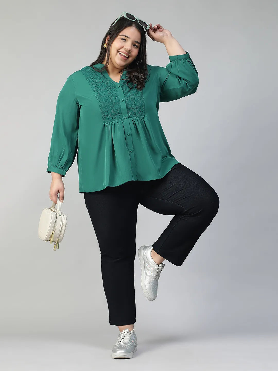 A smiling woman wearing a green blouse with lace detailing and puffed sleeves, posing confidently with her hand on her hip.