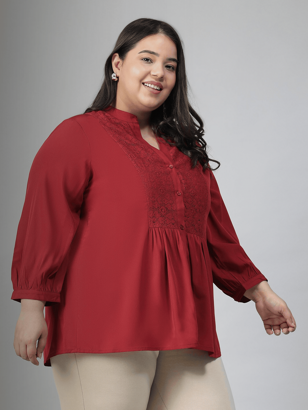 A smiling woman wearing a red blouse with a lace pattern and a high collar, posing confidently with her hand on her hip.