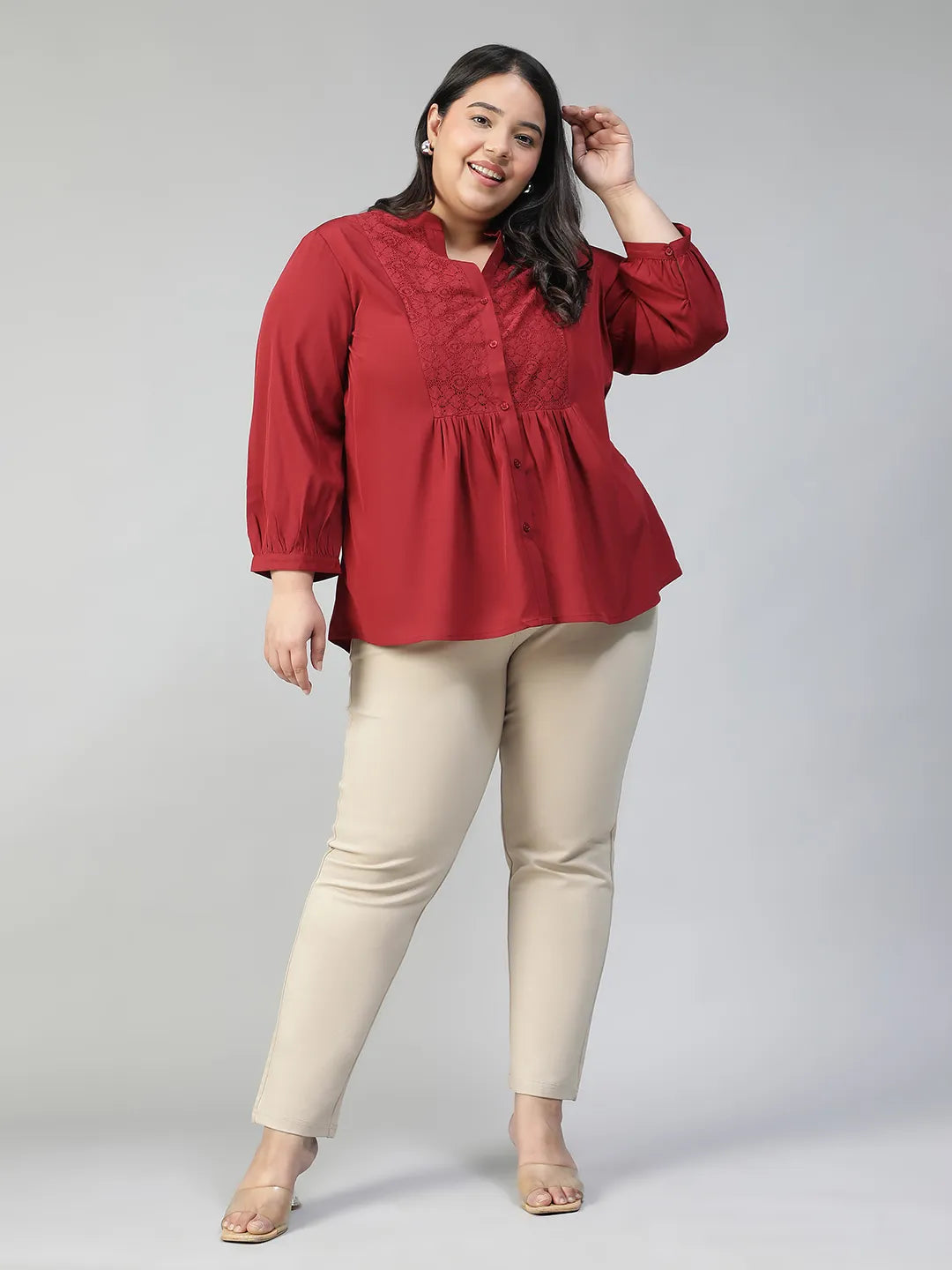 A smiling woman wearing a red blouse with a lace pattern and a high collar, posing confidently with her hand on her hip.