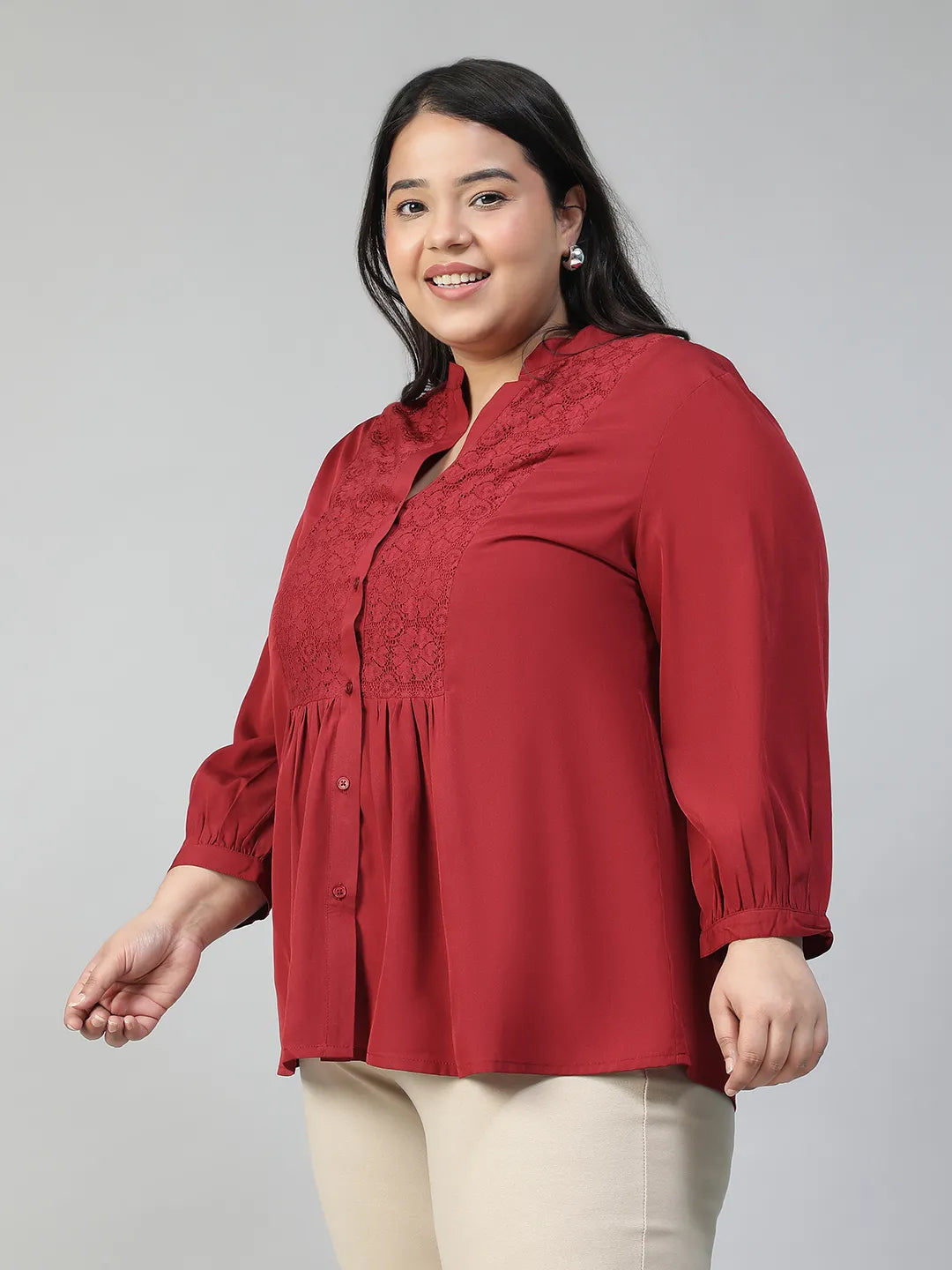 A smiling woman wearing a red blouse with a lace pattern and a high collar, posing confidently with her hand on her hip.