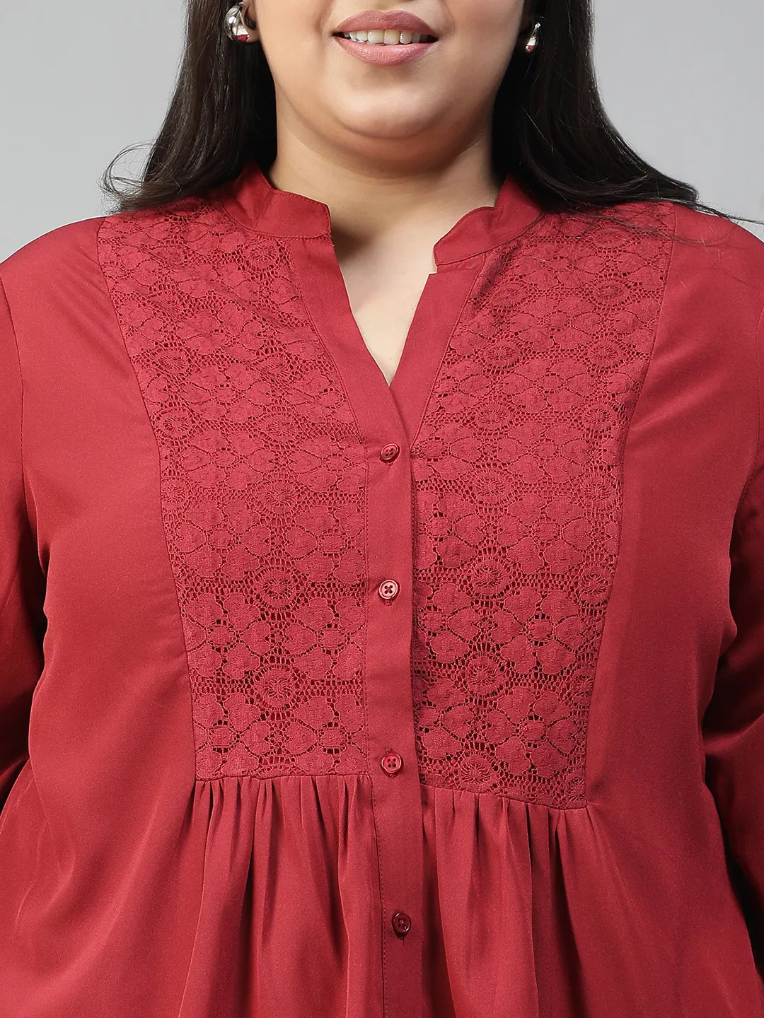 A smiling woman wearing a red blouse with a lace pattern and a high collar, posing confidently with her hand on her hip.
