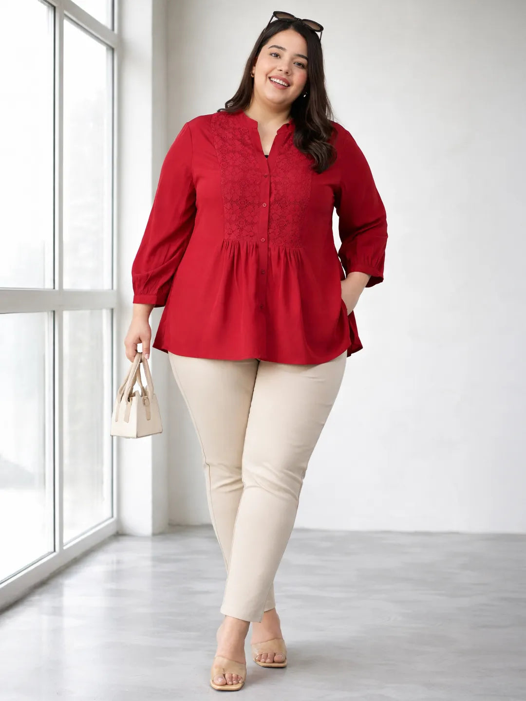 A smiling woman wearing a red blouse with a lace pattern and a high collar, posing confidently with her hand on her hip.