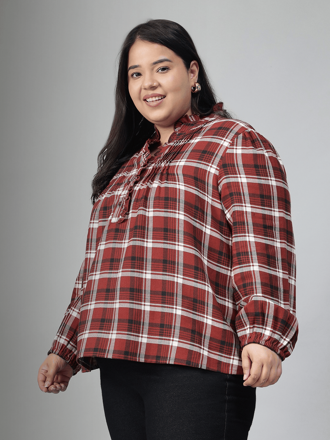 A woman with long dark hair is smiling and posing for the camera, wearing a red and white plaid shirt.