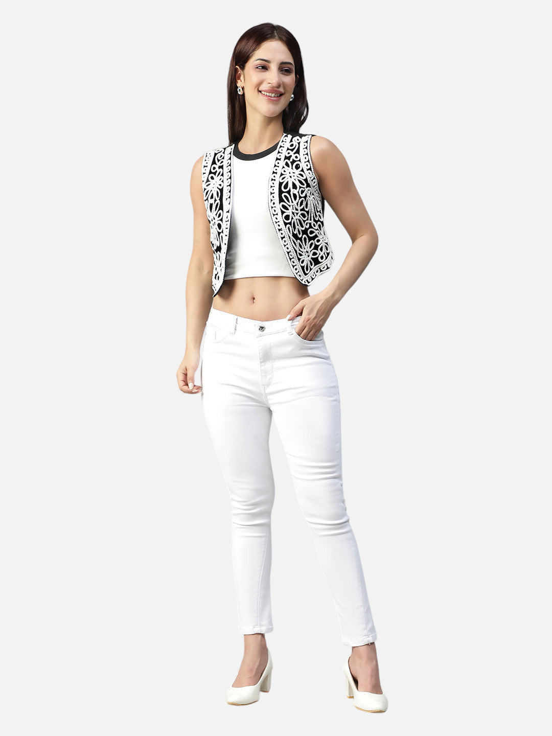 Woman in white cropped top, black and white embroidered vest, white skinny jeans, and heels