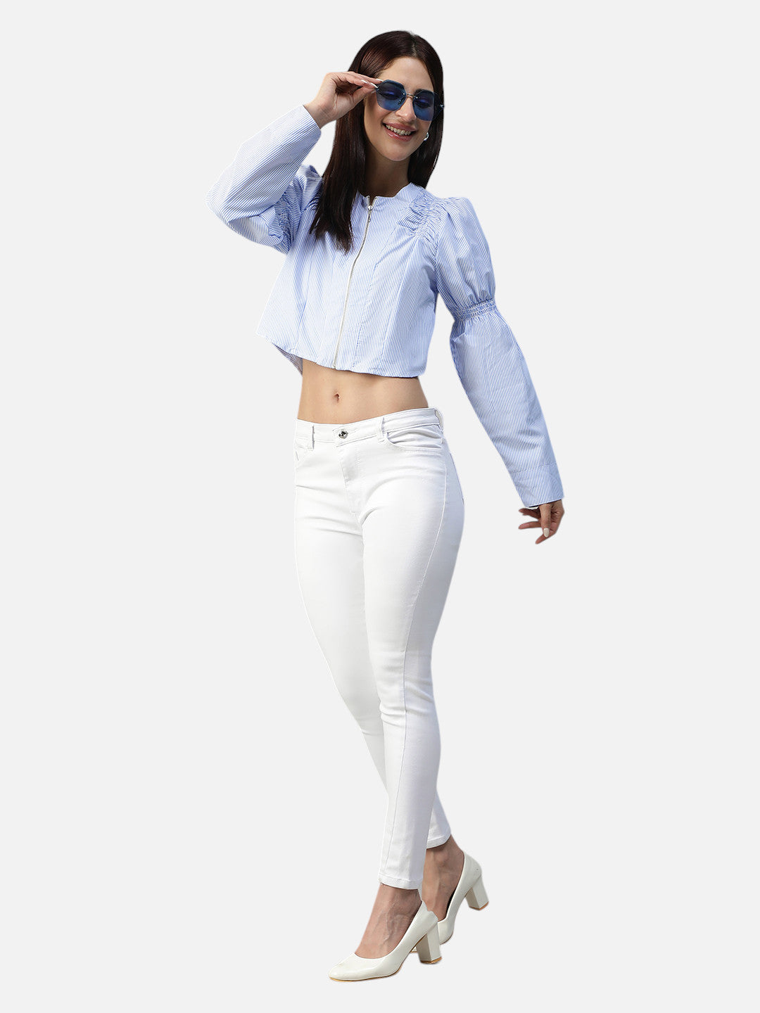 Woman in blue striped crop top and white skinny jeans with white heels on plain background