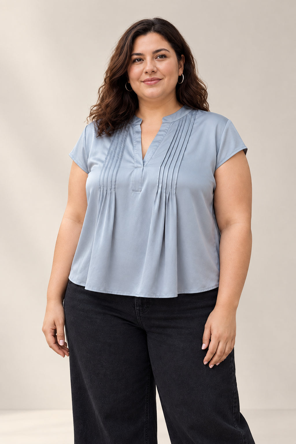 A woman wearing a light blue-gray short-sleeve blouse with pleated detailing on the chest and black pants, standing outdoors on a paved walkway with trees, water, and a railing in the background.