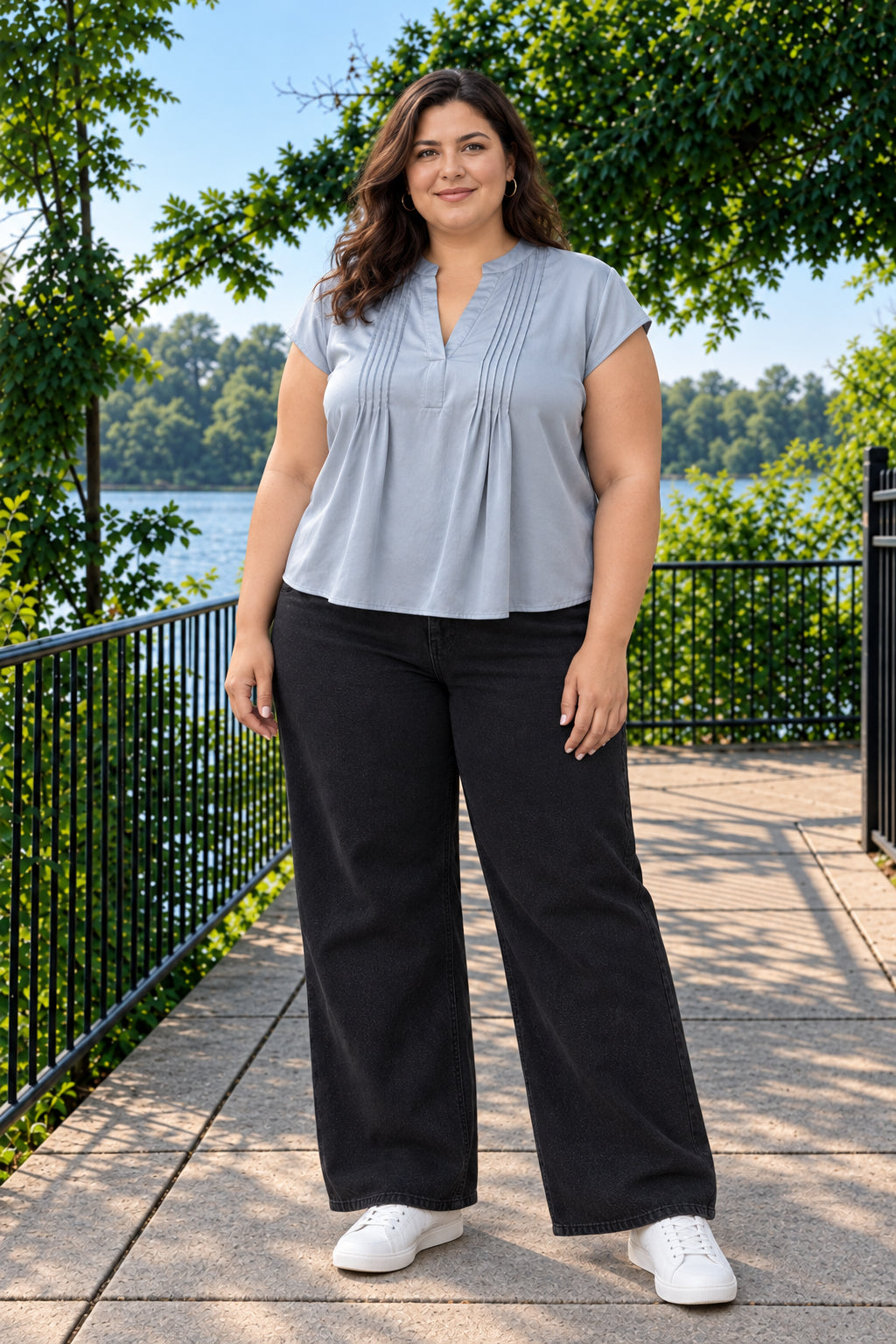 A woman stands outdoors on a paved walkway by a lake, wearing a light blue short-sleeve blouse with pleated detailing and black wide-leg pants, paired with white sneakers.