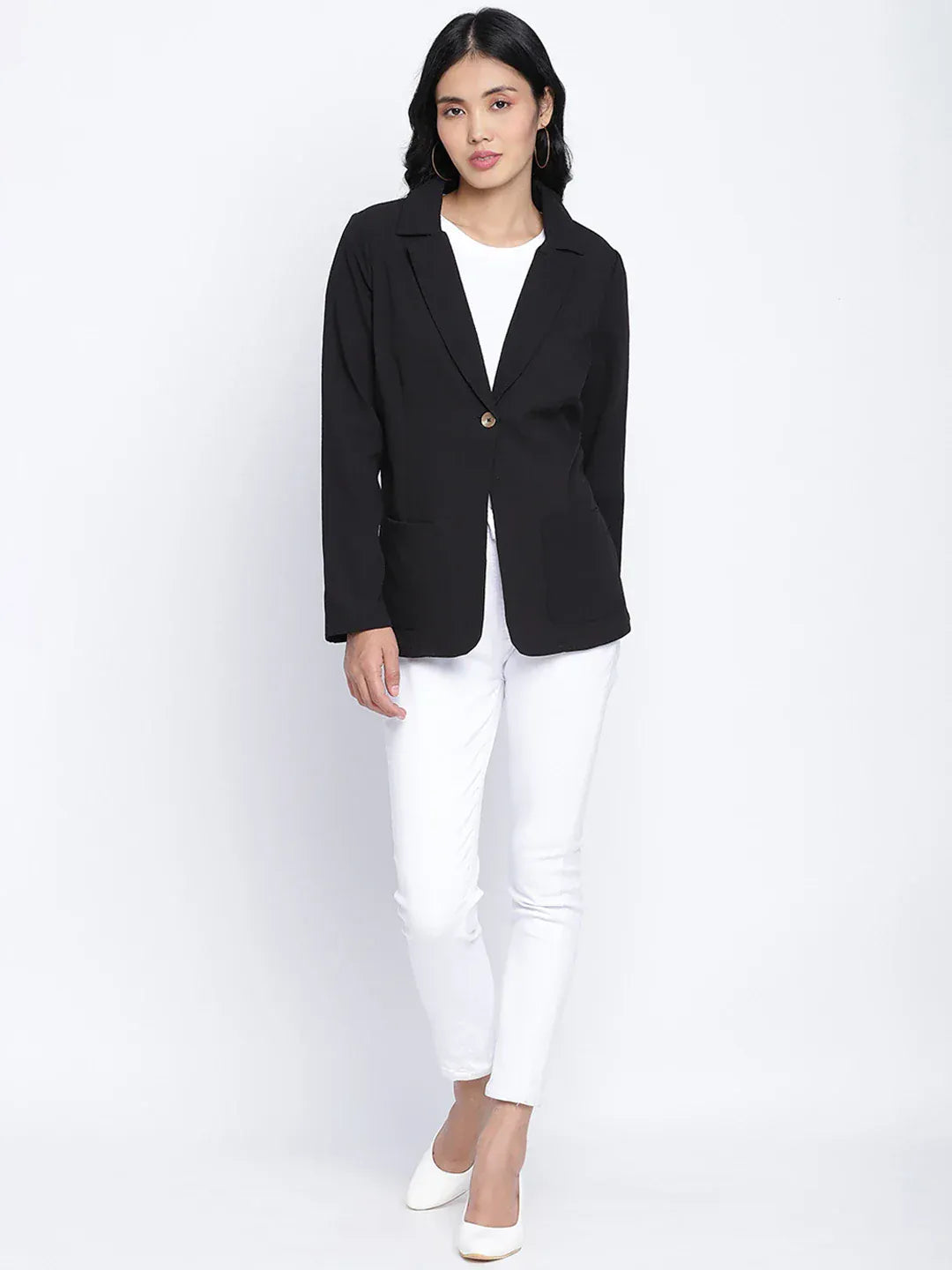 A woman wearing a black blazer, white pants, and white heels, with sunglasses on her head, posing against a white background.