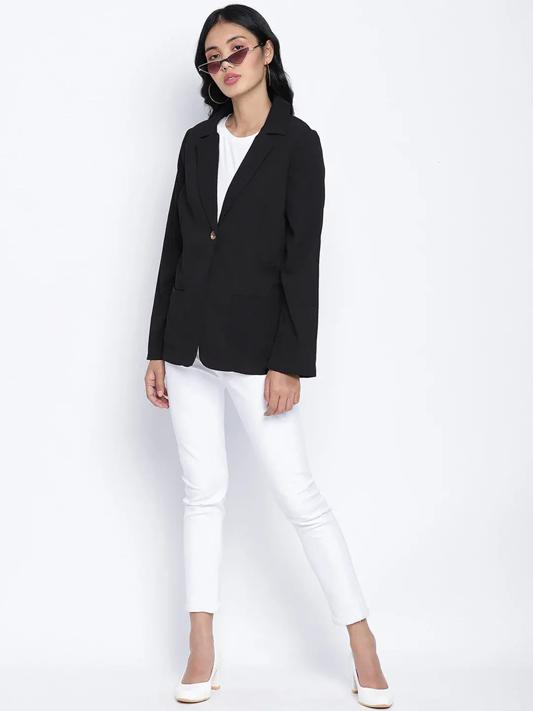 A woman wearing a black blazer, white pants, and white heels, with sunglasses on her head, posing against a white background.