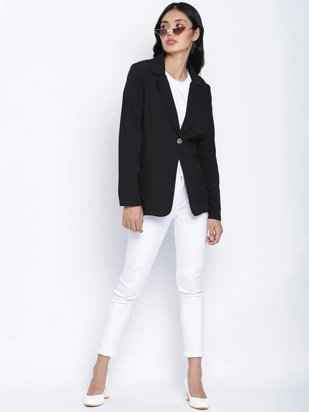 A woman wearing a black blazer, white pants, and white heels, with sunglasses on her head, posing against a white background.