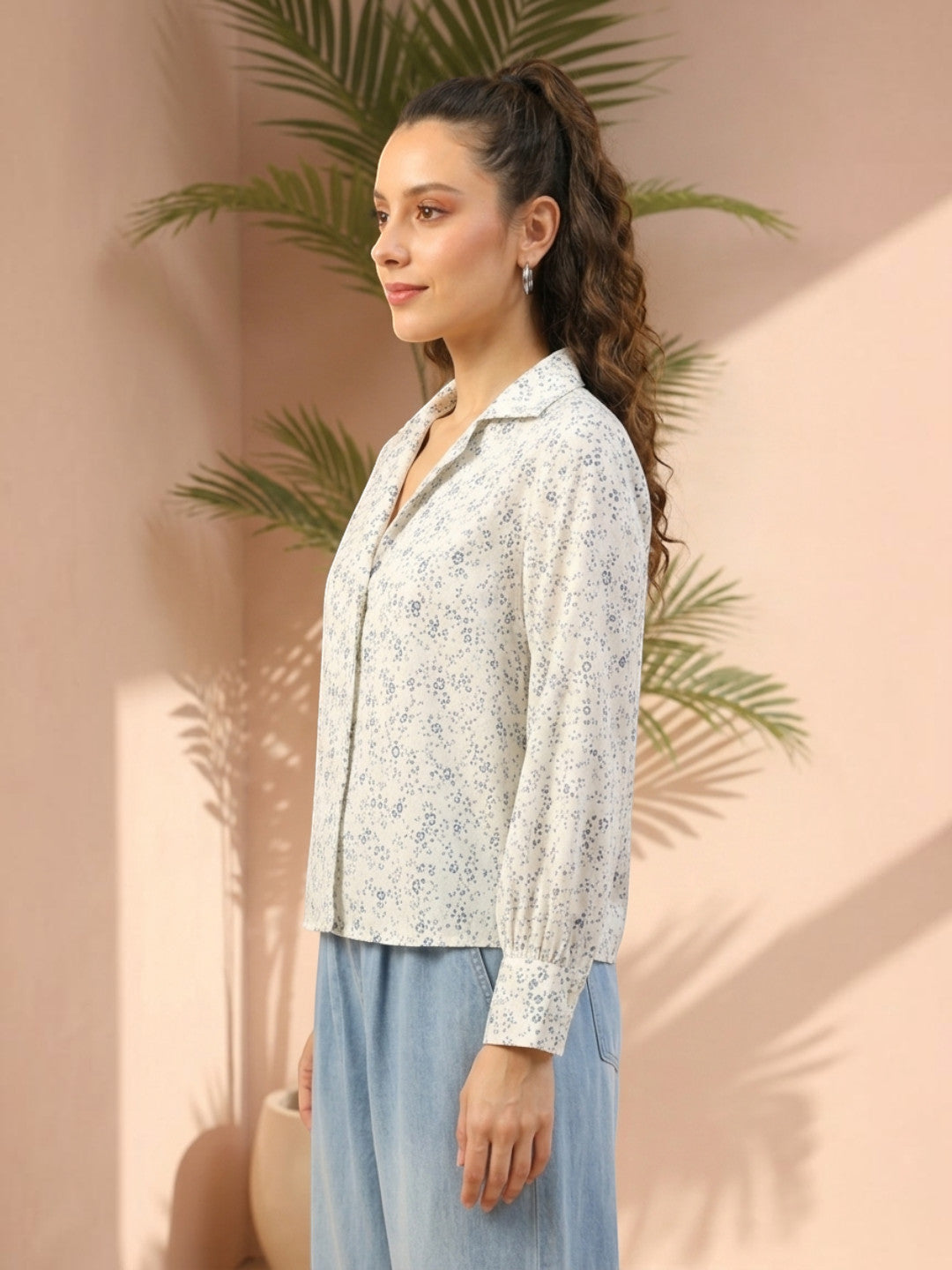 Woman wearing floral print blouse with long sleeves, paired with light blue pants, against pink wall with palm leaves