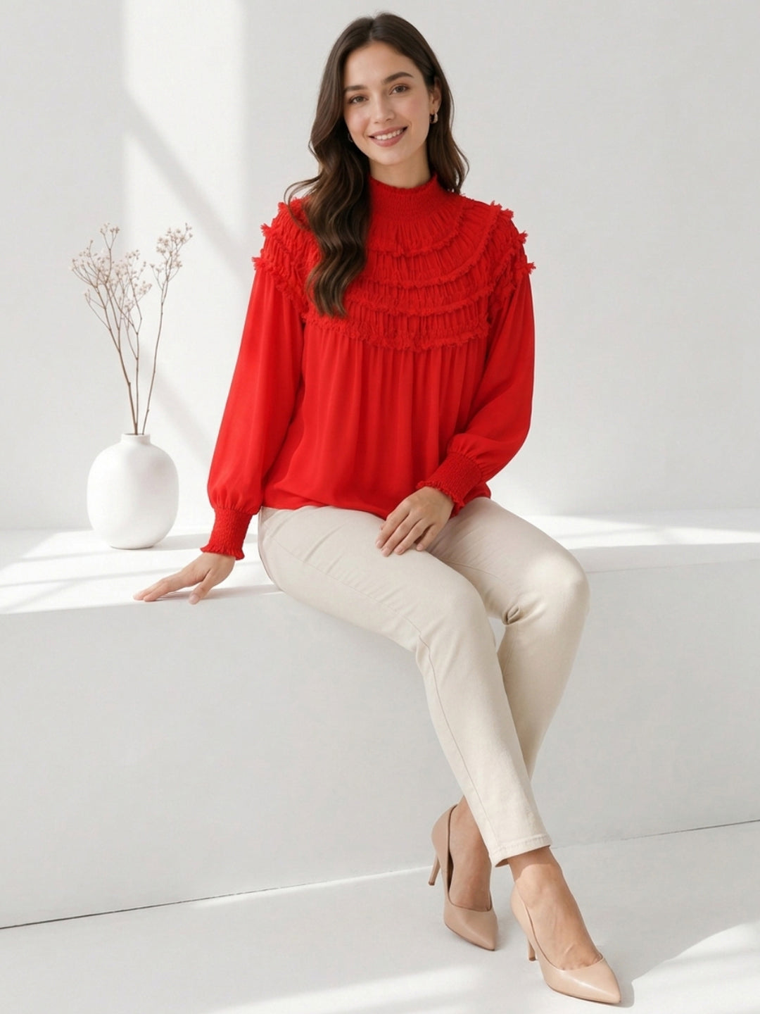 Woman in red ruffled blouse with long sleeves, paired with beige pants and heels, sitting on white bench