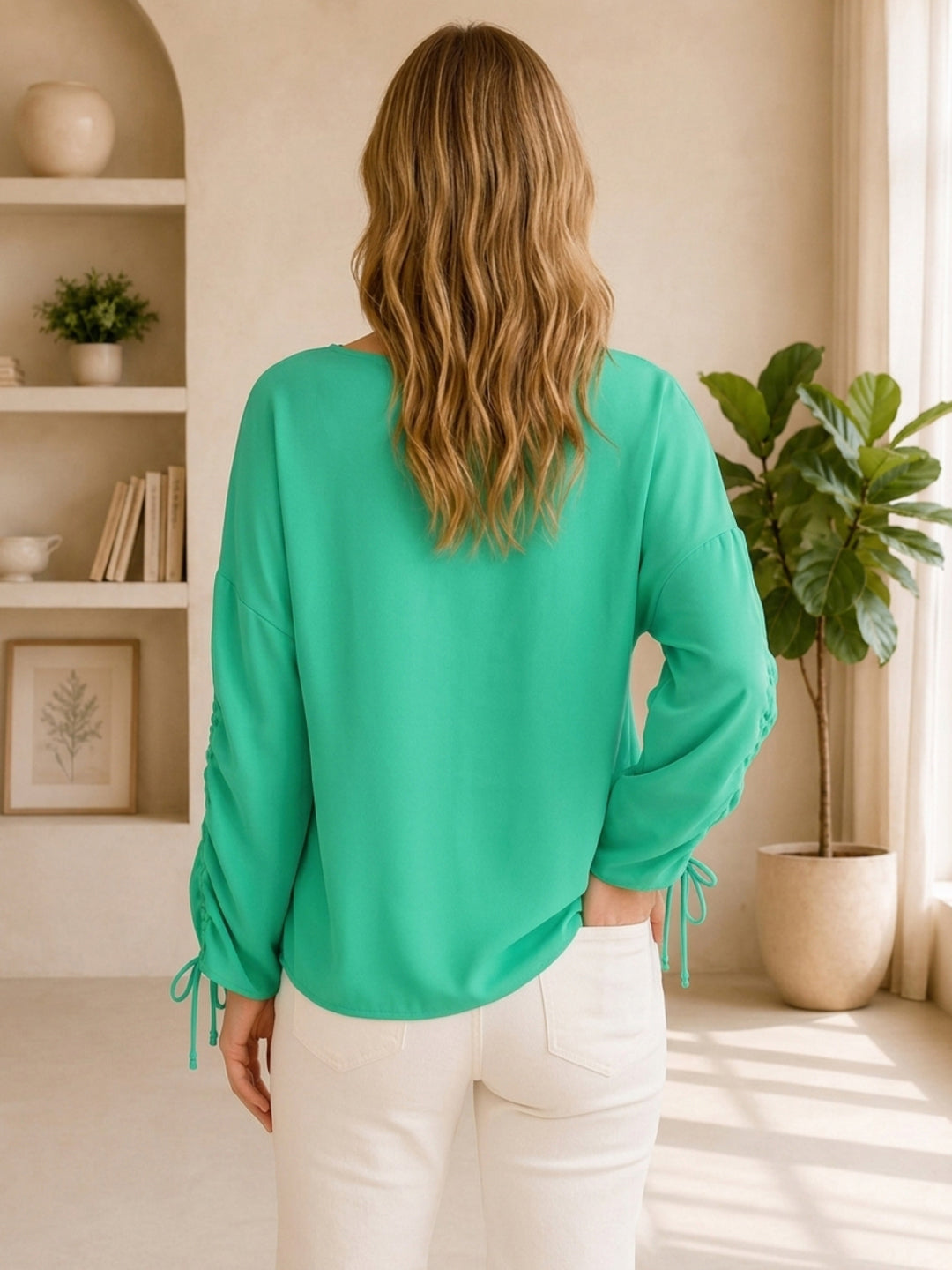 Woman wearing green long sleeve blouse with drawstring sleeves, paired with white jeans, back view