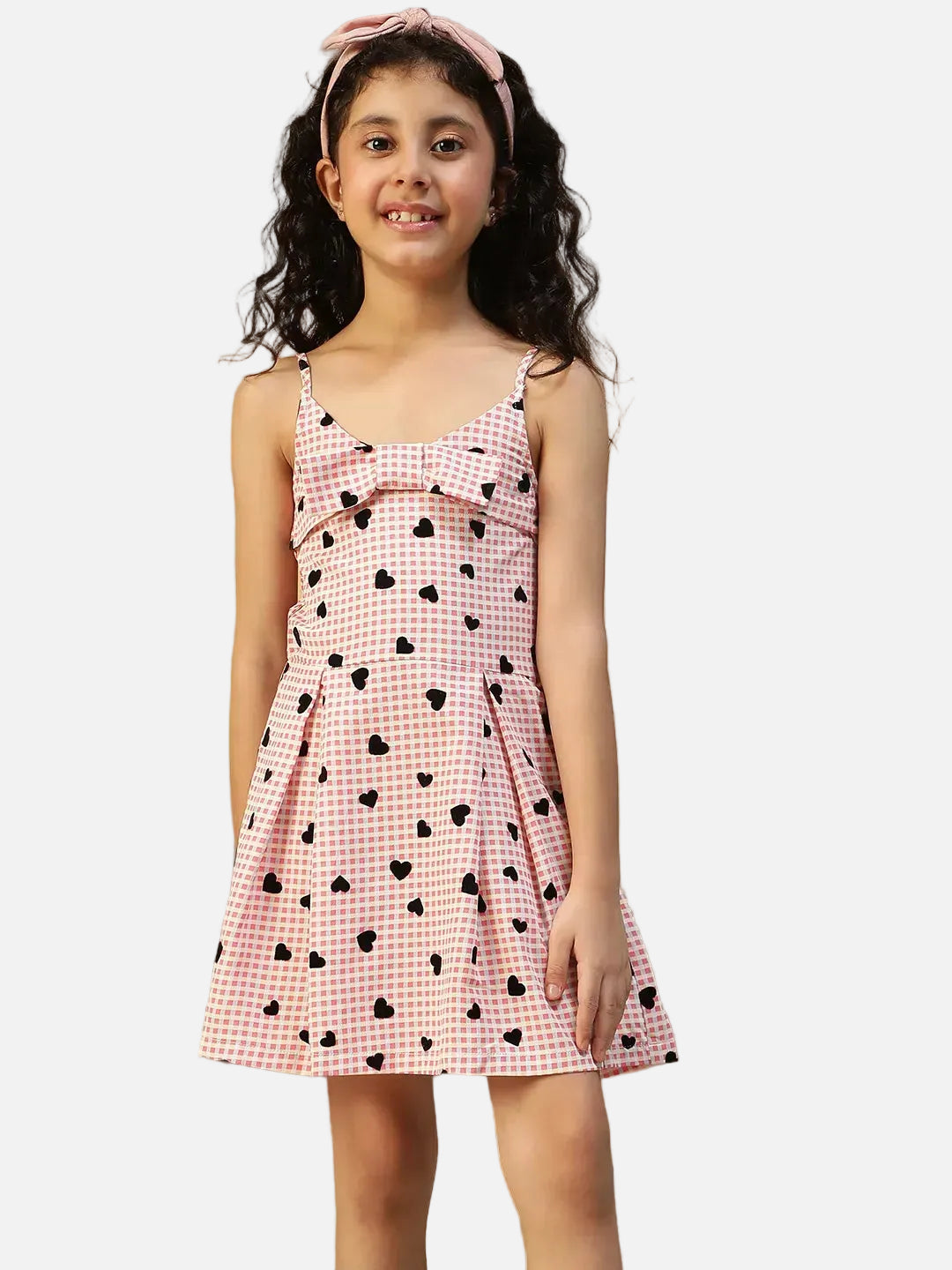 A young girl wearing a pink and white checkered dress with heart patterns stands in front of a plant, smiling at the camera.