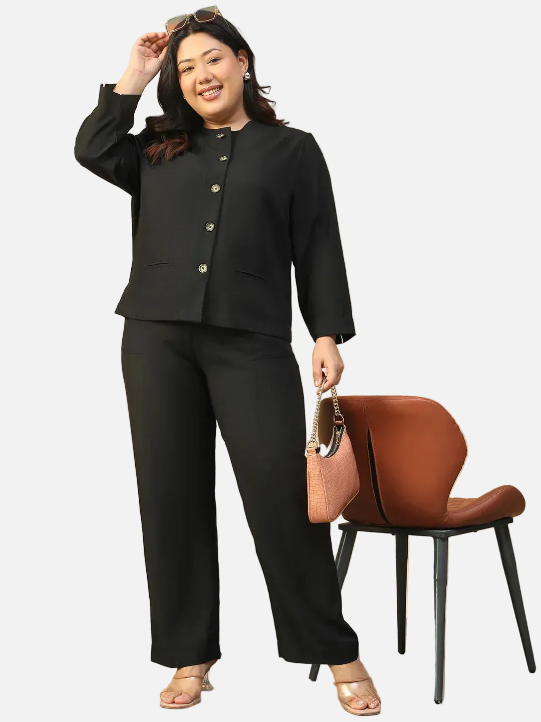 A woman in a black suit and heels is standing next to a brown leather chair, smiling and holding a small purse.