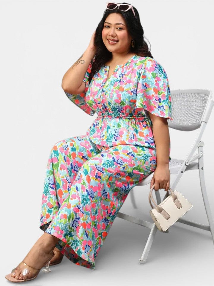 A woman wearing a colorful floral print jumpsuit is sitting on a white folding chair, smiling and holding a small purse.