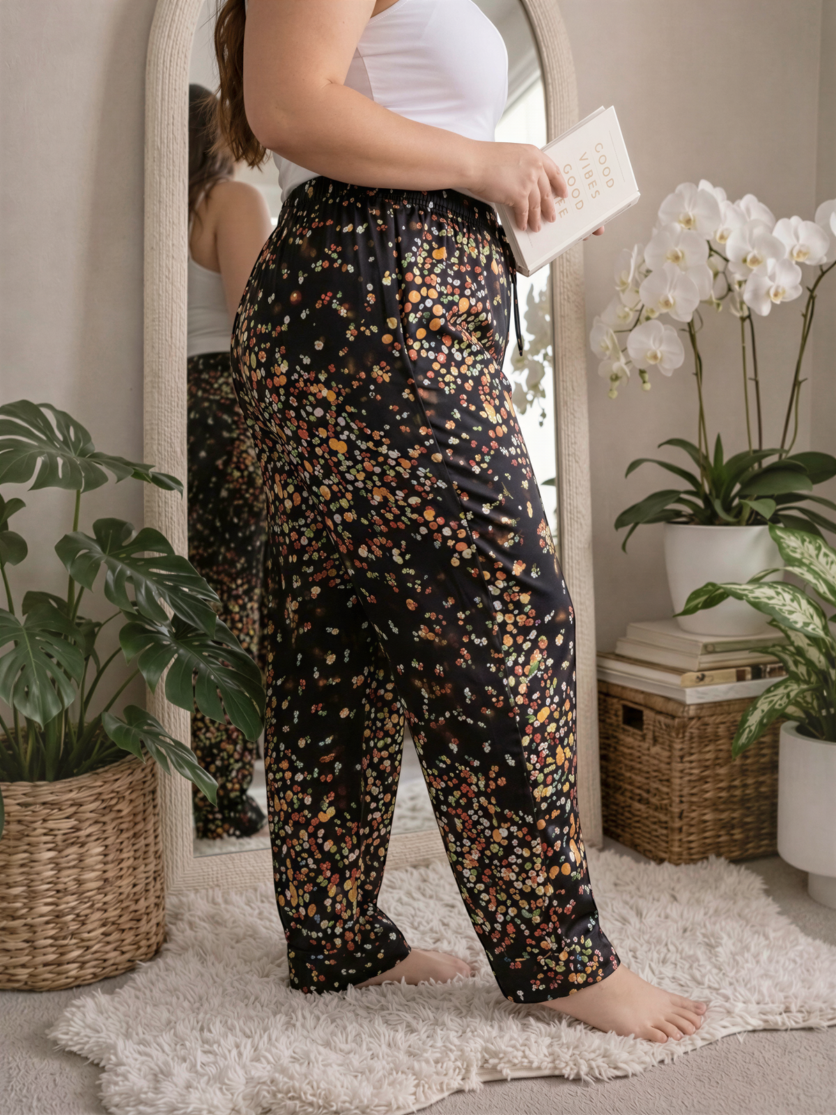 A woman stands in front of a mirror, holding a book, with a floral print skirt and a white top. The room is decorated with potted plants and a white rug.