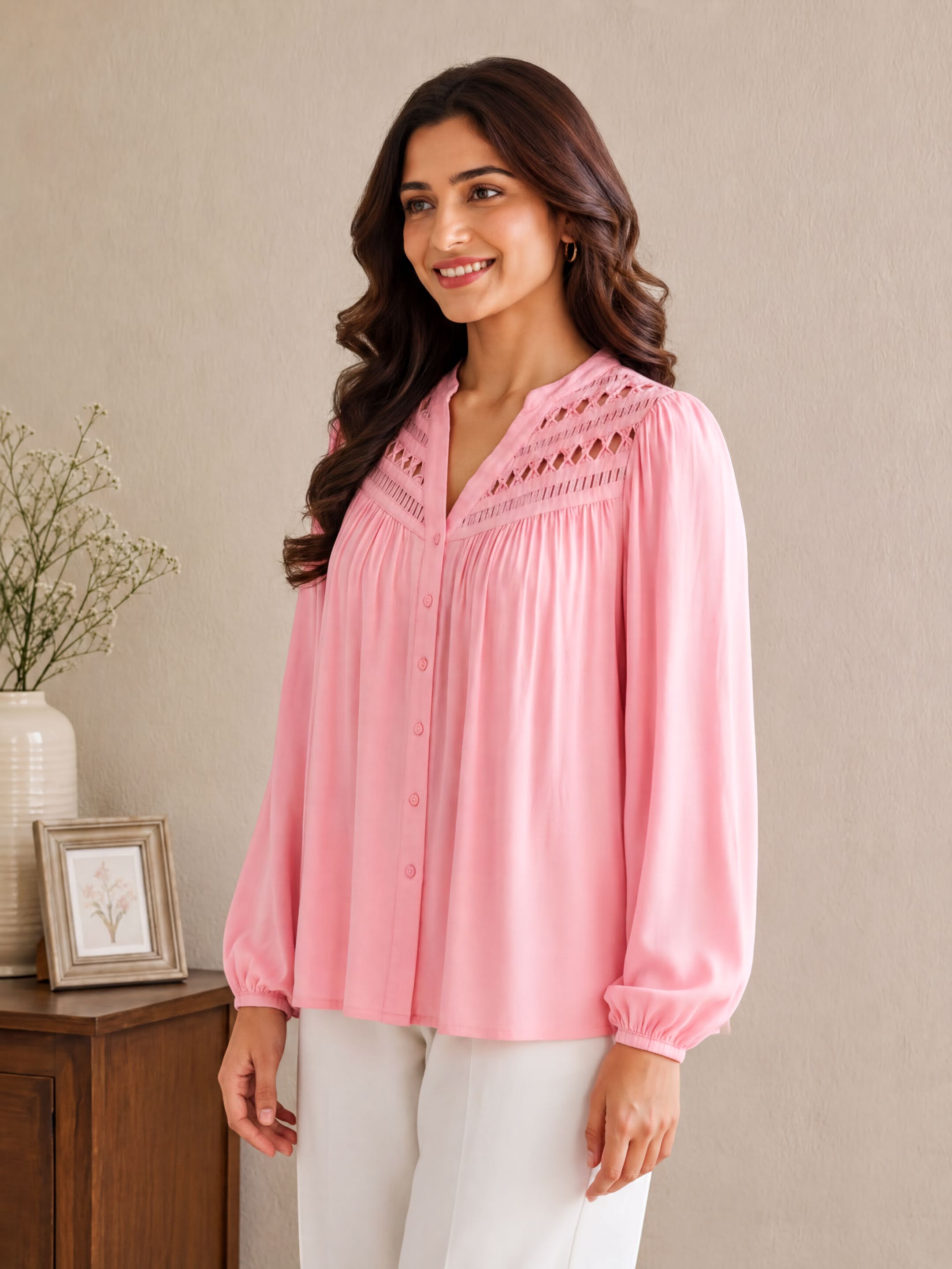 A woman wearing a pink blouse with a ruched collar and long sleeves stands in front of a wooden cabinet with a picture frame on it.