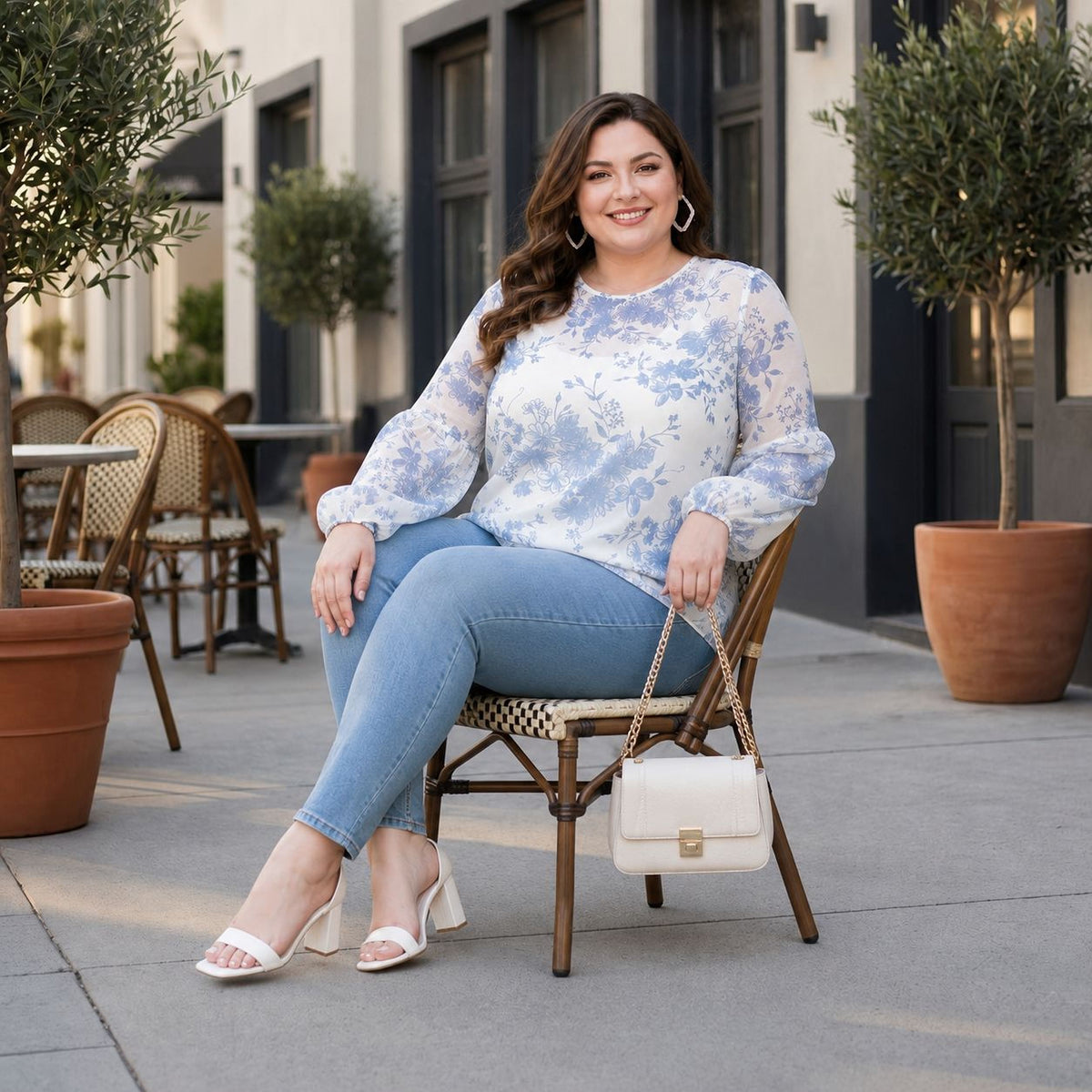 A woman wearing a white blouse with blue floral patterns, blue jeans, and white heels is sitting on a chair outdoors, smiling and holding a white purse.