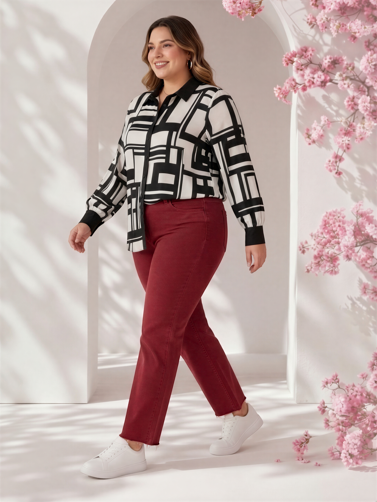 A woman wearing a black and white patterned shirt and red pants is standing in front of a white archway with pink flowers.