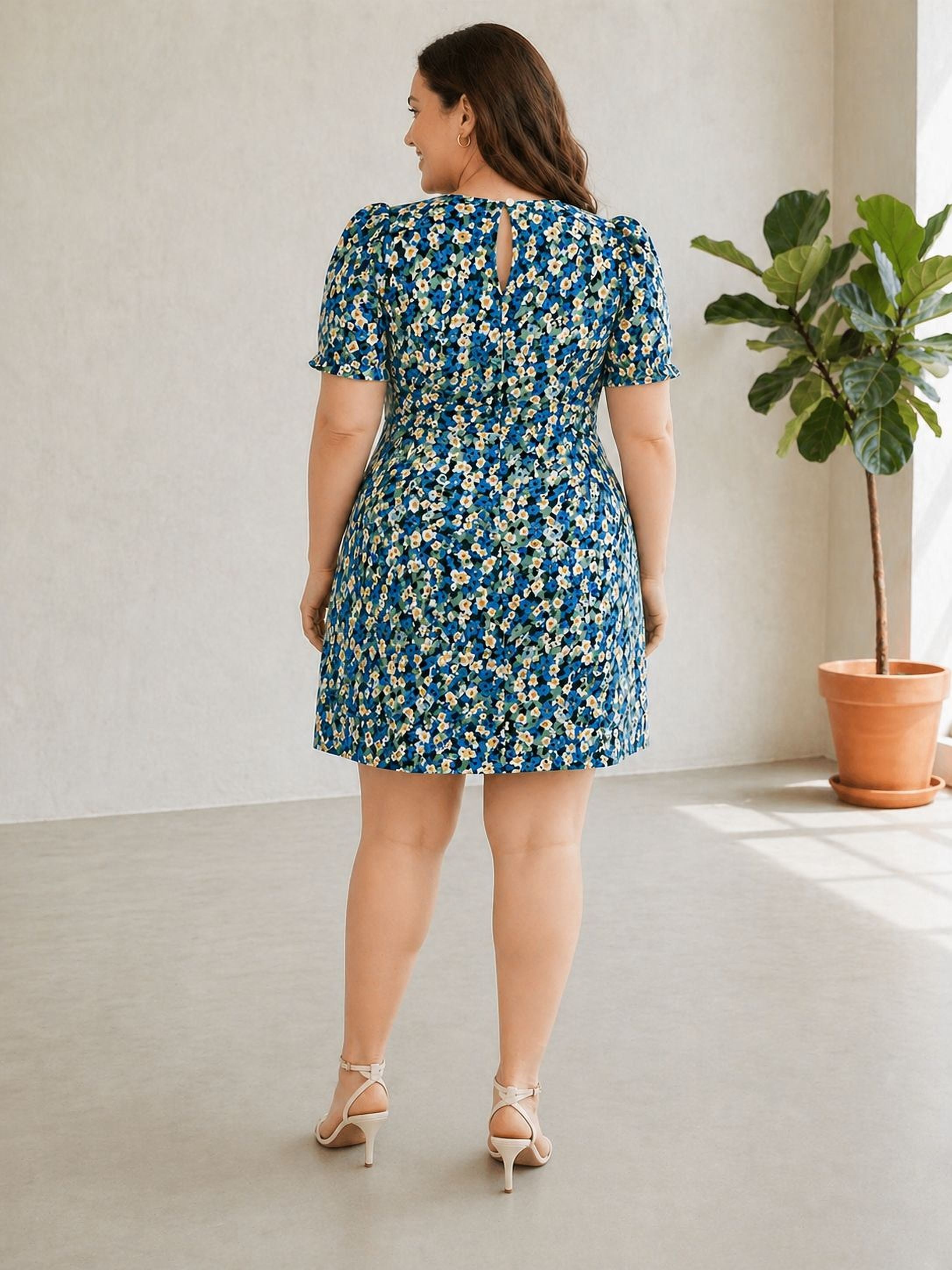 A woman wearing a blue and white floral print dress stands in a room with a potted plant to her right.