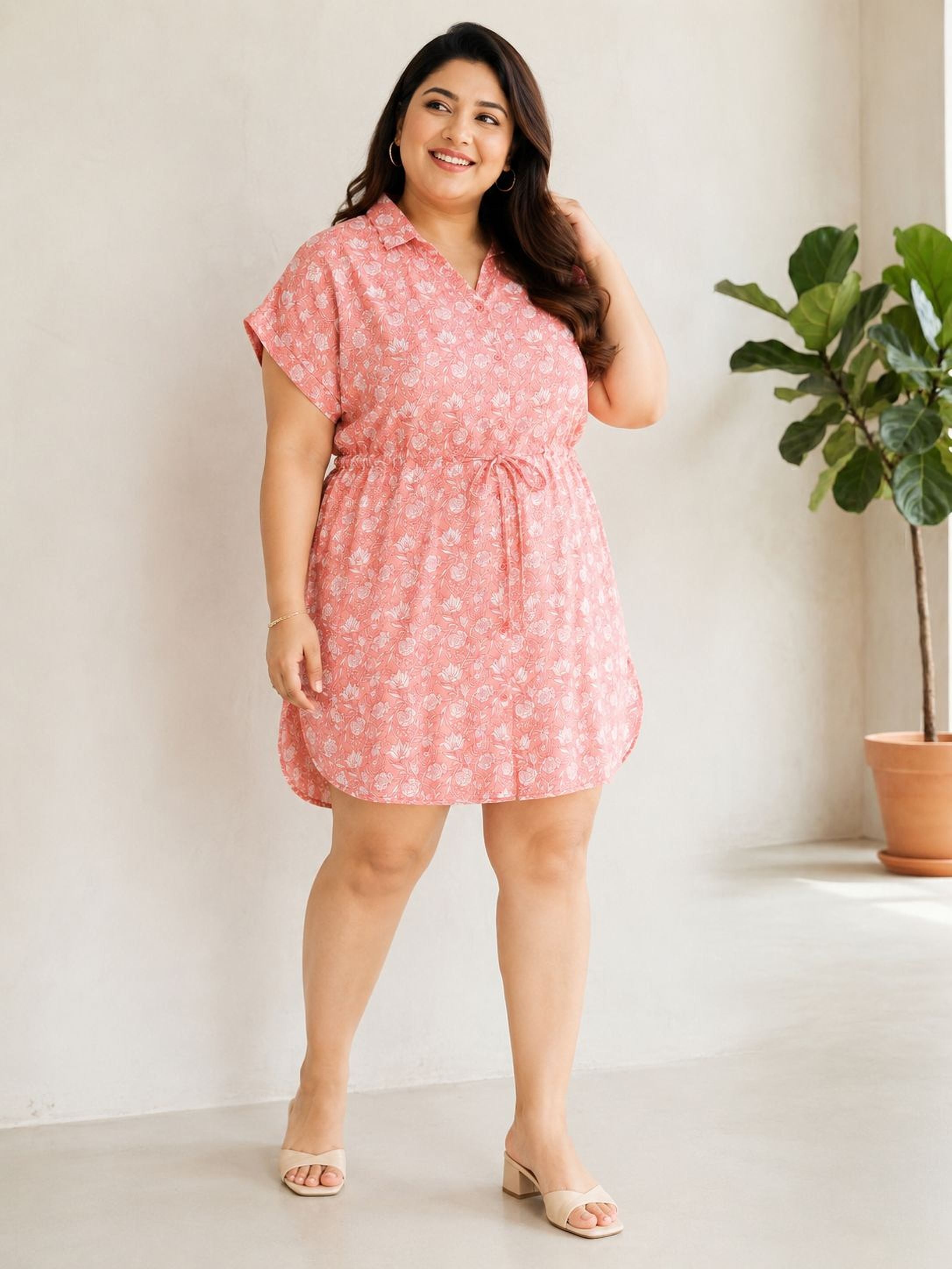 A woman wearing a pink floral dress stands in a room with a potted plant in the background.
