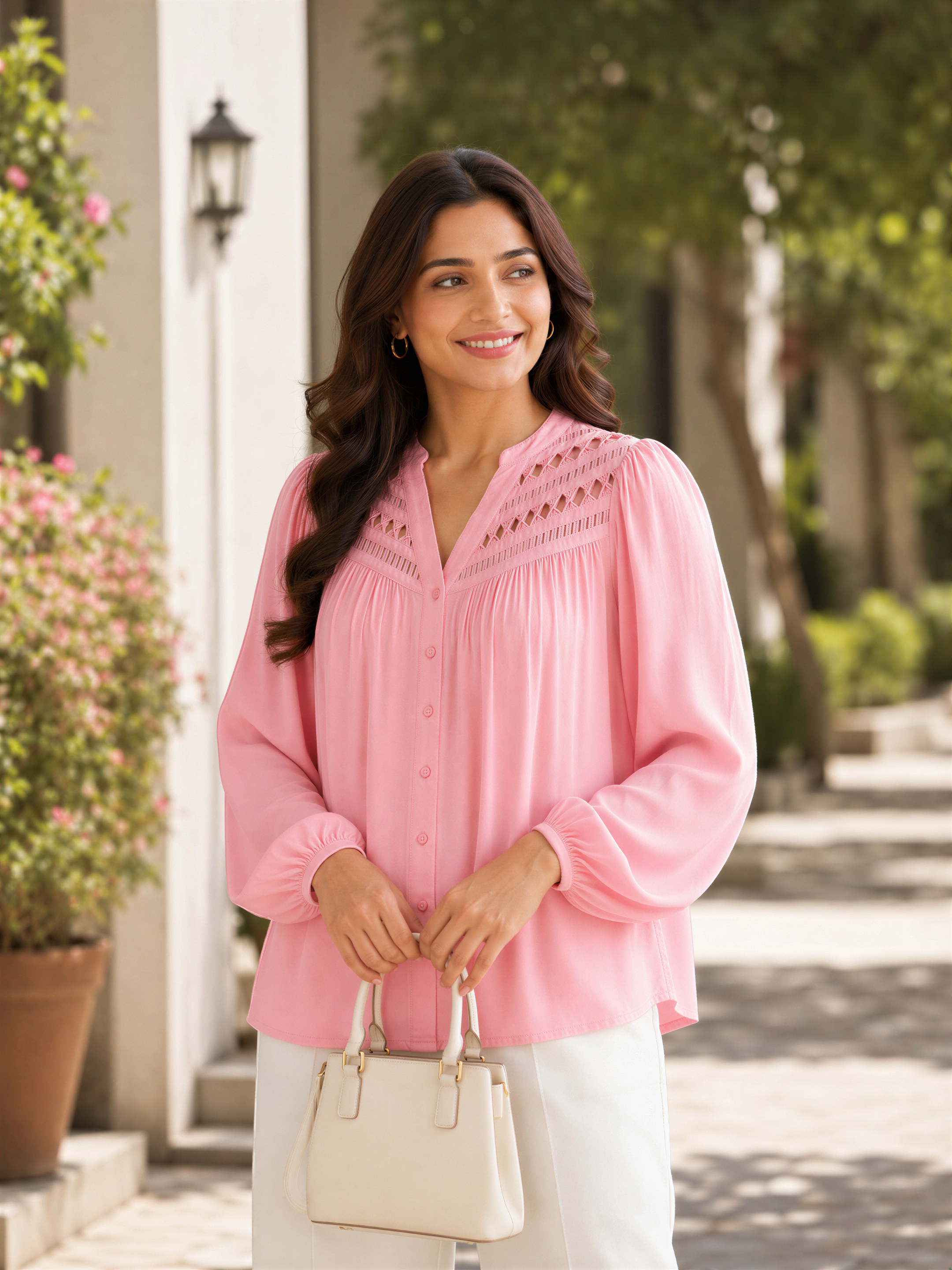 A woman in a pink blouse and white pants stands on a sidewalk, smiling and holding a beige purse.