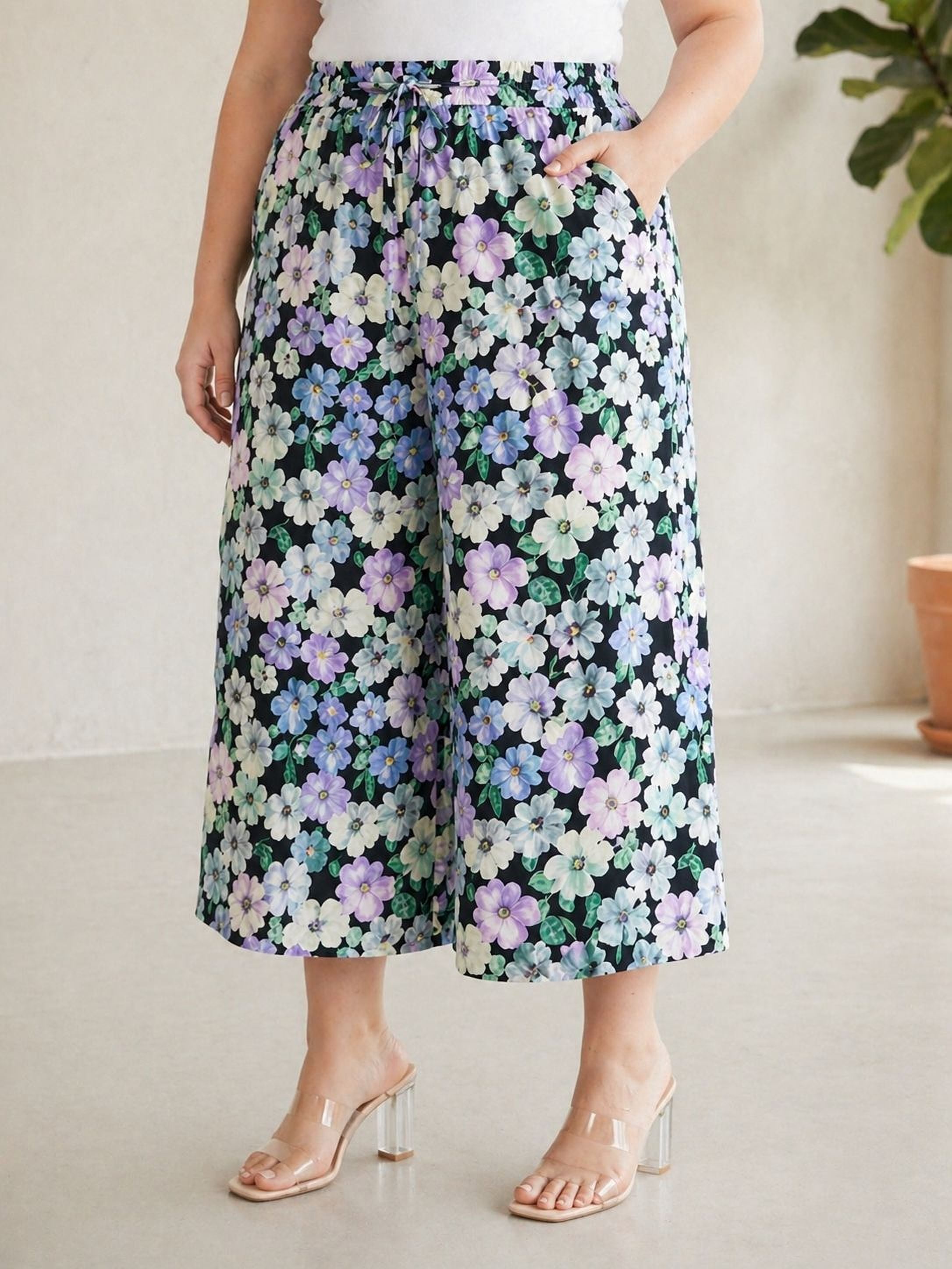A woman wearing a floral print skirt and clear high heels stands in a room with a potted plant in the background.