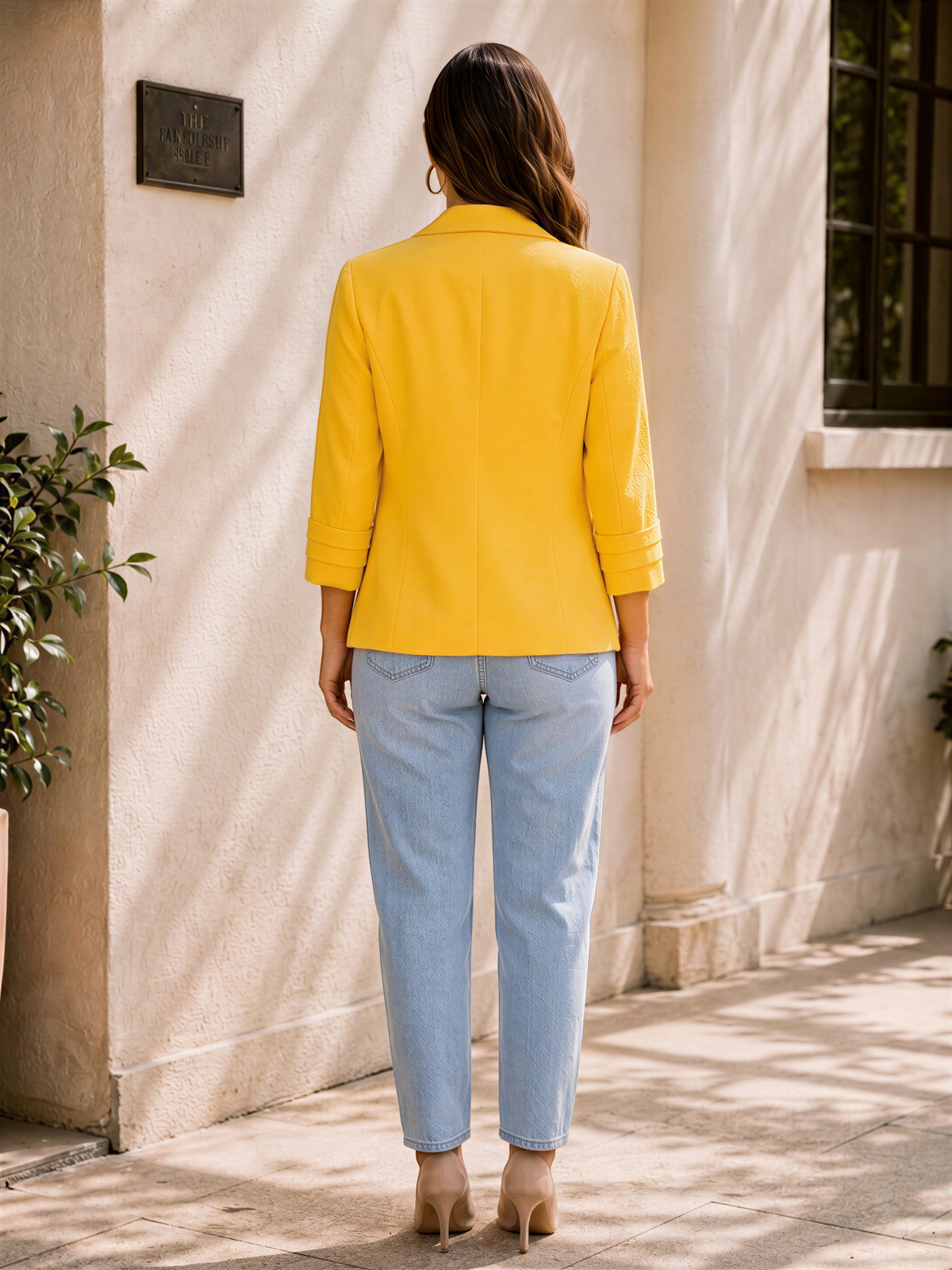 A woman wearing a yellow blazer and blue jeans stands in front of a building, facing away from the camera.