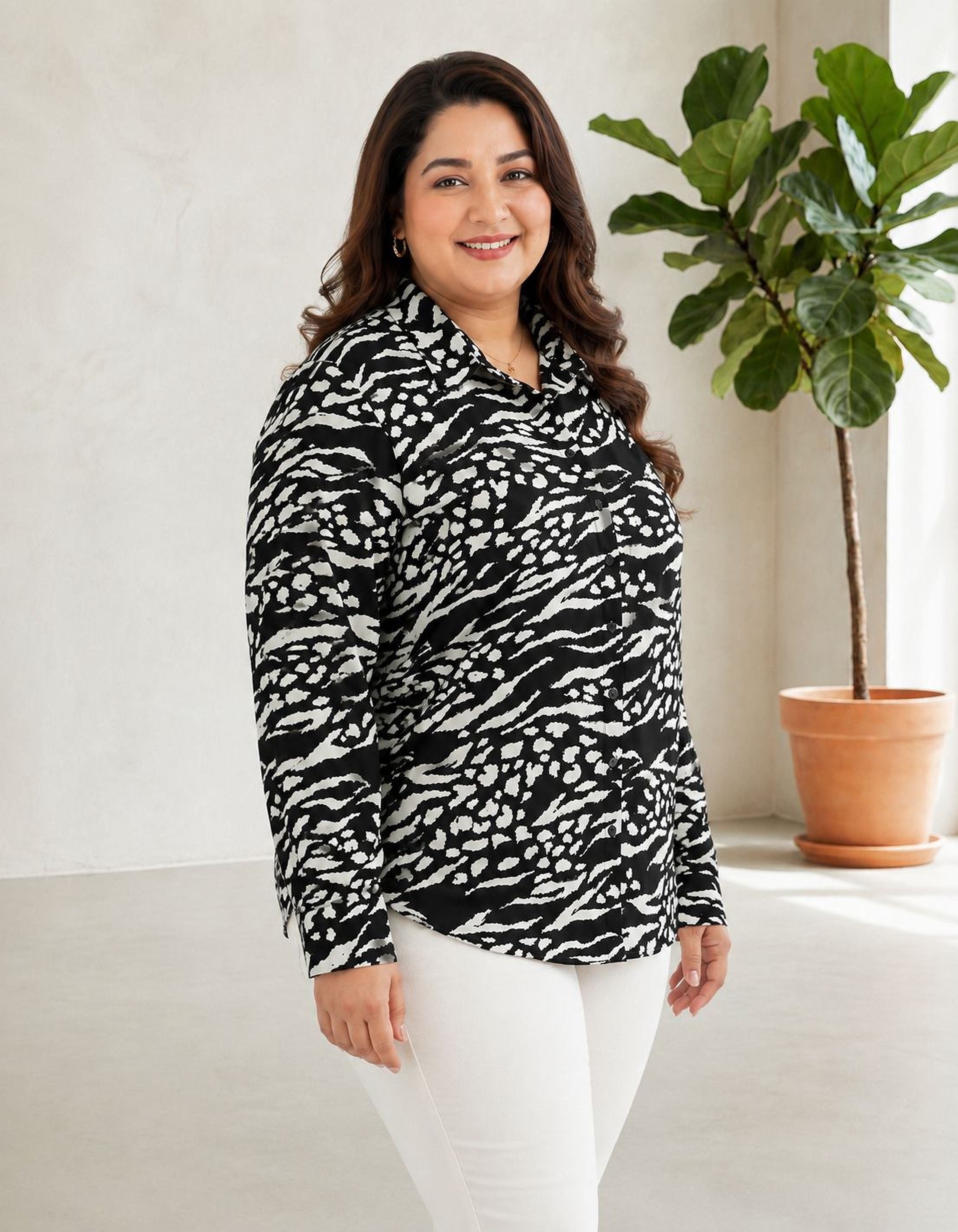 A woman with long brown hair is standing in a room, smiling at the camera. She is wearing a black and white patterned blouse and white pants. In the background, there is a potted plant.